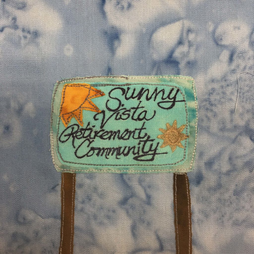 This quilt block shows a teal sign supported by two brown poles. The sign has a large sun in yellow-orange fabric in the top left corner, and another sun, more gold, in the bottom right corner like a badge. The writing on the sign is black and is a combination of cursive and print, that reads, "Sunny Vista Retirement Community."
