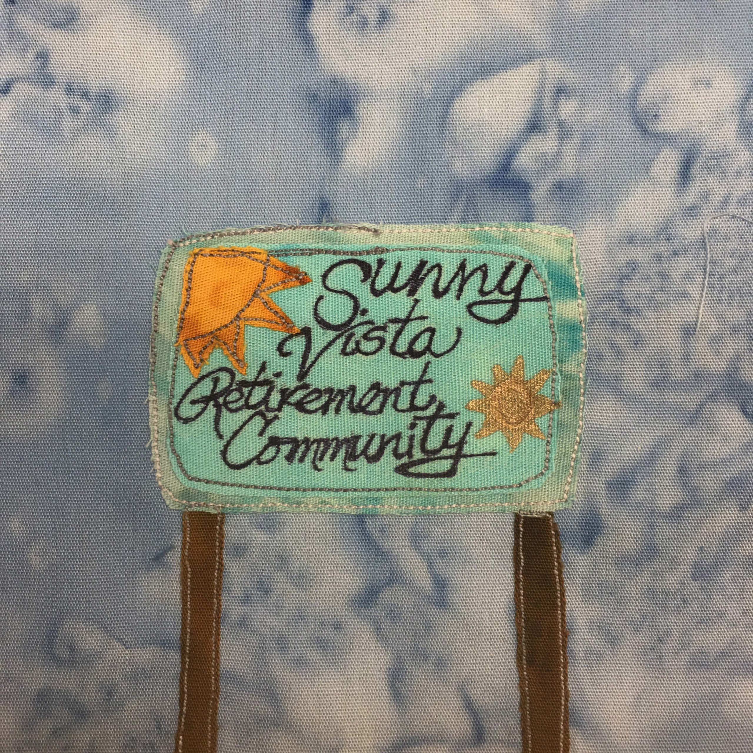 This quilt block shows a teal sign supported by two brown poles. The sign has a large sun in yellow-orange fabric in the top left corner, and another sun, more gold, in the bottom right corner like a badge. The writing on the sign is black and is a combination of cursive and print, that reads, "Sunny Vista Retirement Community."