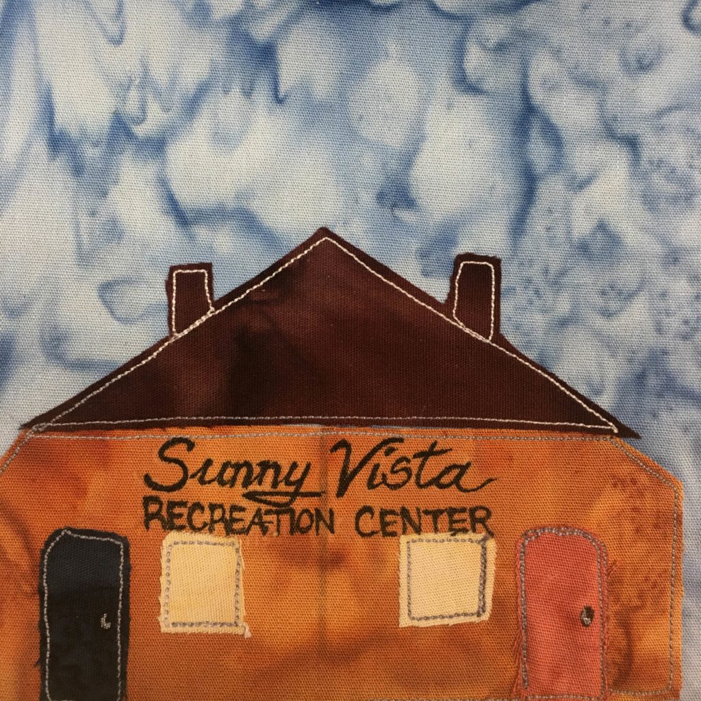 This quilt block shows a wide orange building, almost as wide as the block itself. The roof is a triangle of very dark brown with two chimneys, individually outlined in stitching, poking out on either side. Below the triangular roof is a large orange piece of fabric making the main body of the building. On the front are the words in black pen, 