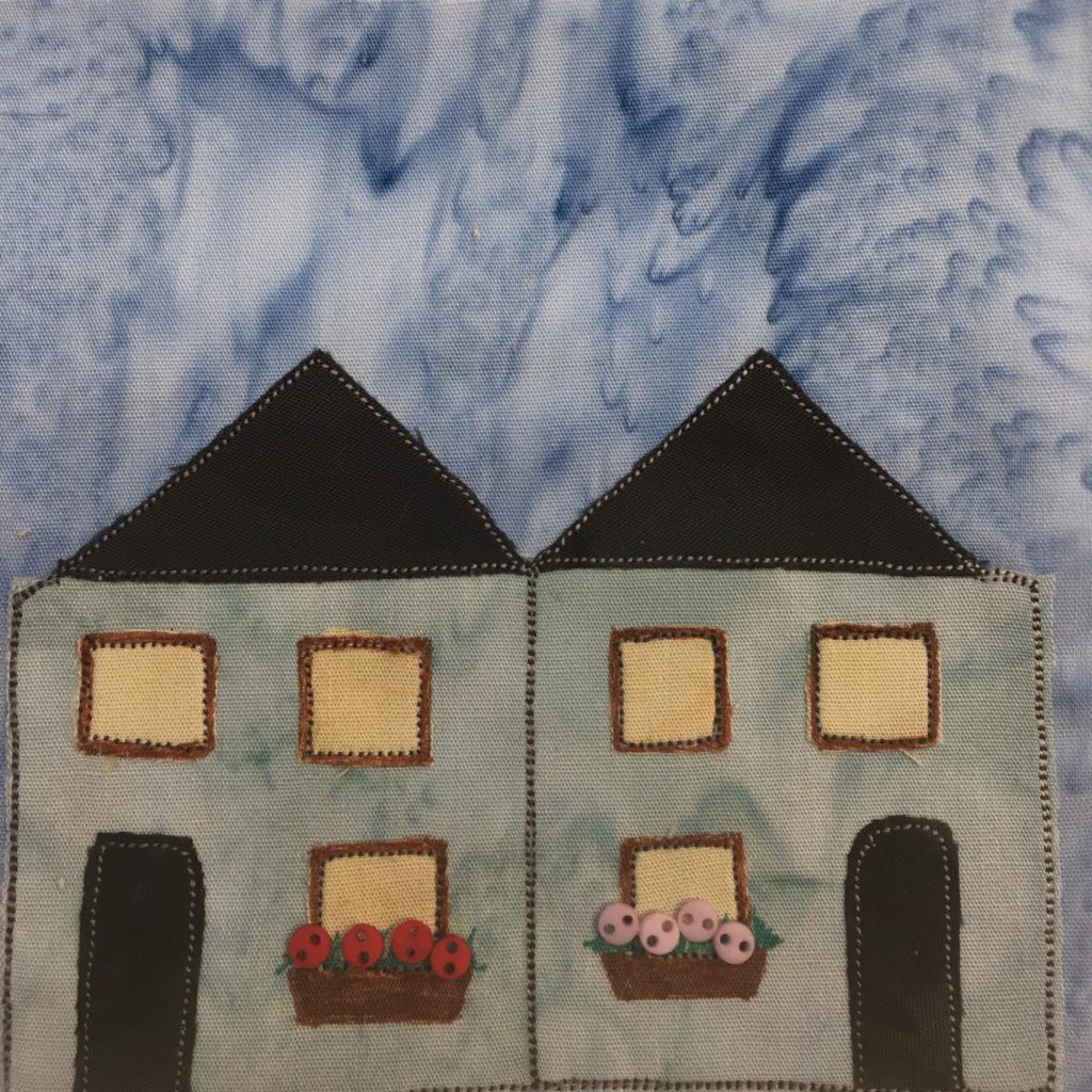 This quilt block shows two building side by side. They are really one building made up of two townhomes. Each townhome has a black triangular roof. Below that is a beige-teal square building. The fronts of the townhomes are mirror images of each other. Each one has two upper floor square yellow windows (bordered by brown thread, and one lower floor yellow square window. The lower windows have flowerbeds in front of them, with one having red flowers (tiny two-hole buttons), and the other having pink flowers. Next to each window is a black rounded door.
