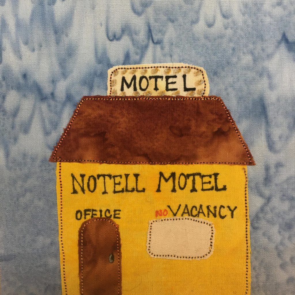 This quilt block shows a yellow building with a brown roof (and door). Above the roof is a sign with lights around it (indicated by marks of pen) that says, "Motel" The roof is in a trapezoid shape and is a deep brown fabric. Underneath the roof on the yellow building are the words, "Notell Motel". Above the door it reads, "Office" and above the light tan window it reads, "No Vacancy". Each piece of fabric, including the tan window, is outlined by a duotone black and light tan stitching.