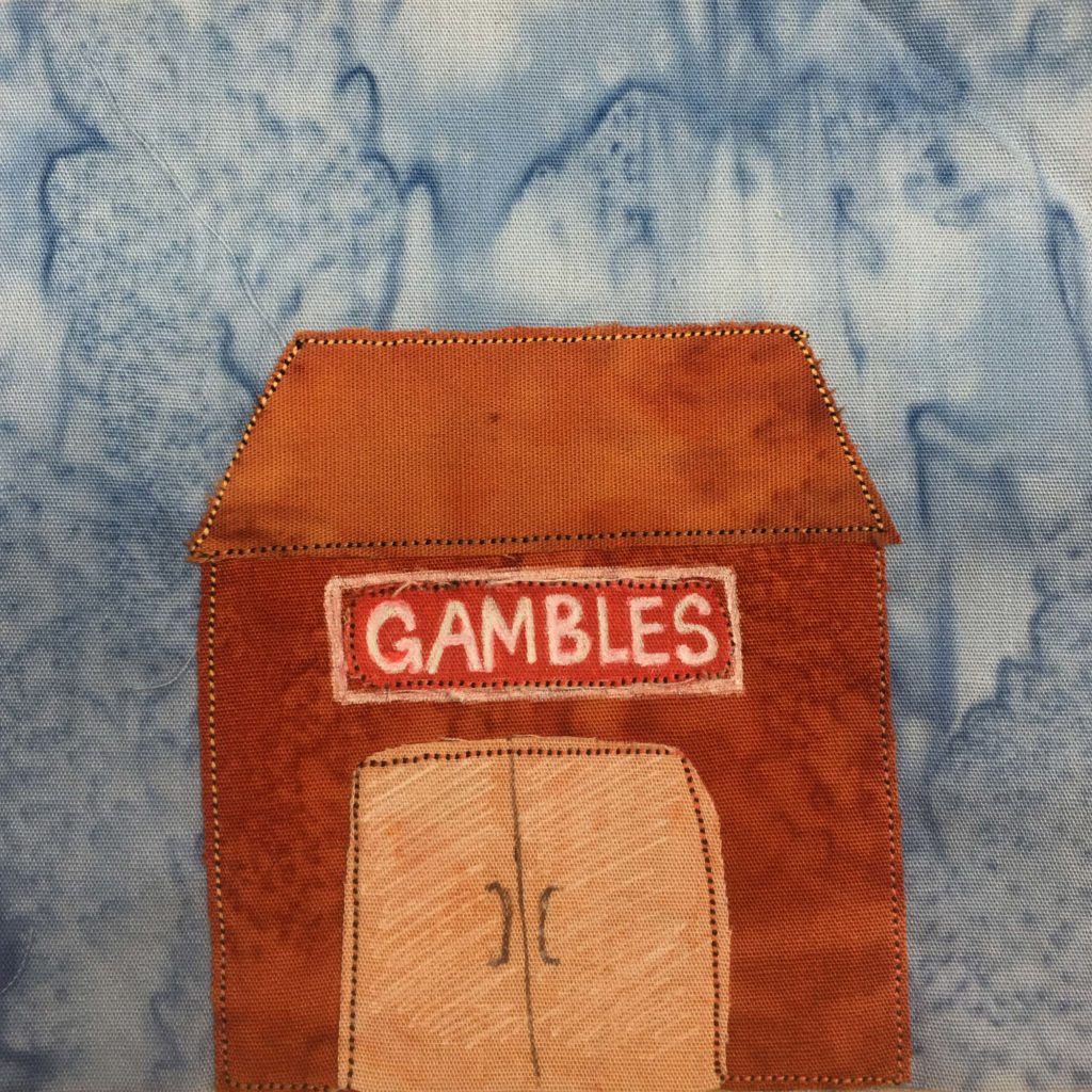 This quilt block shows a very orange building with an orange roof and a light orange glass door (shaded with white pen). The sign above the double doors is red with a white and black border. It reads, "Gambles" in white pen in bold letters across it. All the fabric pieces are outlined by an orange and black stitching.