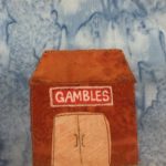This quilt block shows a very orange building with an orange roof and a light orange glass door (shaded with white pen). The sign above the double doors is red with a white and black border. It reads, "Gambles" in white pen in bold letters across it. All the fabric pieces are outlined by an orange and black stitching.