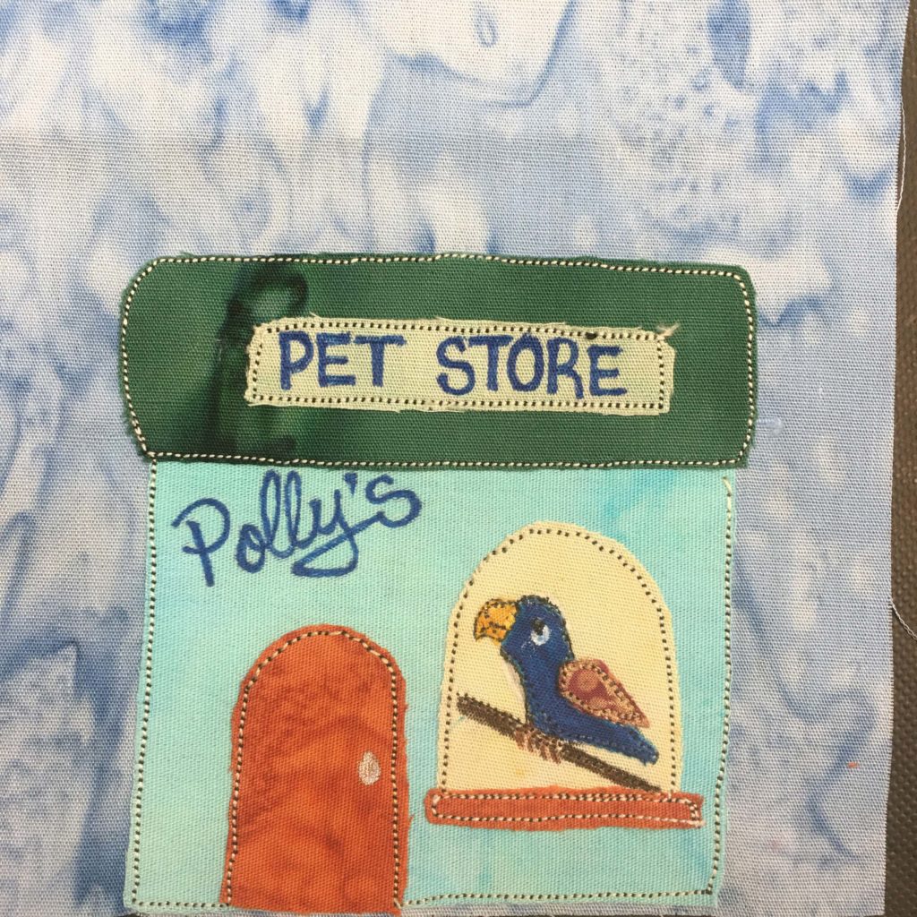 This quilt block shows a very colorful square building. The flat rectangular roof is a deep green fabric with a light green sign in the middle of it with writing in blue pen, "Pet Store." Below that the building is a light teal blue. In the upper left of the building is the word in blue pen, "Polly's" in cursive. On the left is an orange door with a rounded top. To the right is a large light yellow window with an orange trim on the bottom. In the window is a fairly detailed parrot sitting on a perch. The parrot is a deep blue with a yellow beak and light brown wings.
