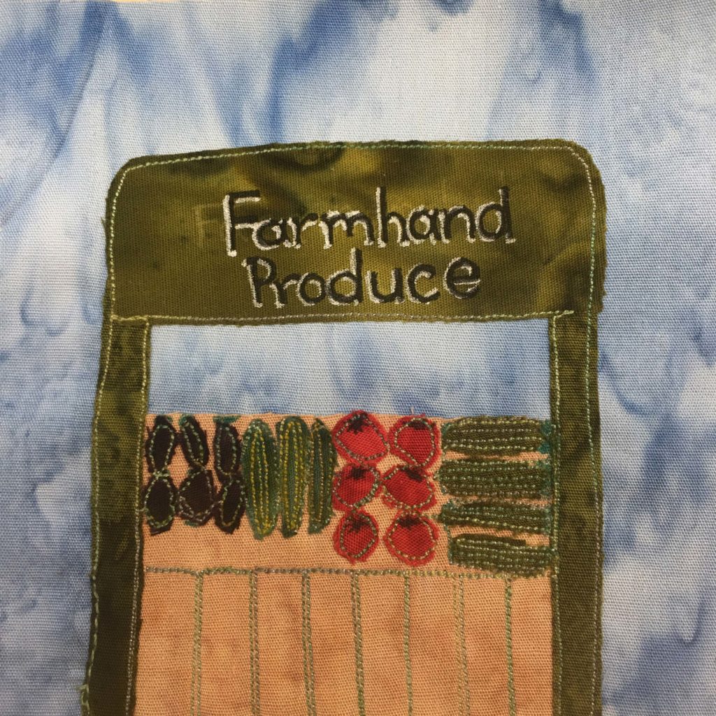 This quilt block shows a green produce stand like you might find on the side of the road. at the top of the stand, going across the green front are the words