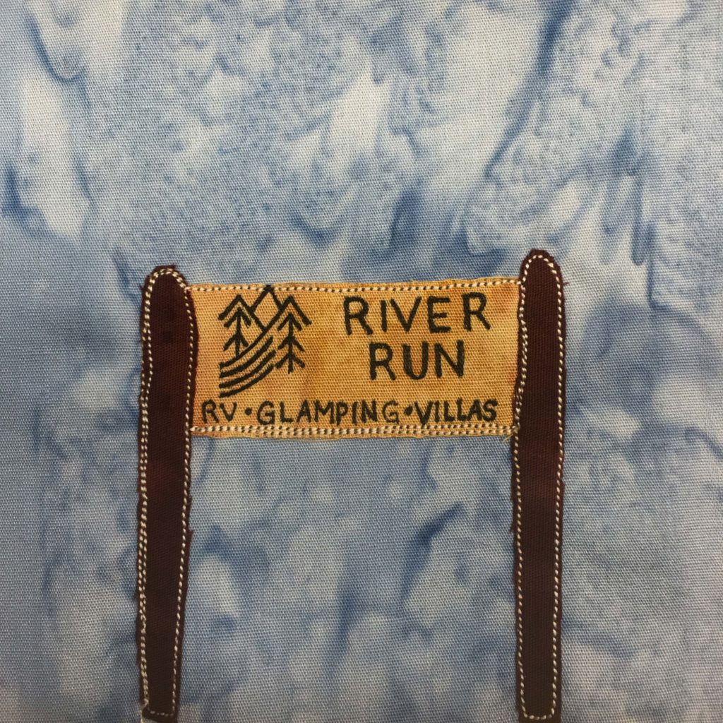 This quilt block shows a sign like you'd see on the side of the road. The sign is between two dark brown poles. The sign itself is a dark yellow with black pen writing on it. There is a logo with mountains, trees, and what looks like a river. Next to the logo in black pen are the words, "River Run." Under both the logo and the larger words are the words, "RV • Glamping • Villas". Each piece of fabric is outlined with a white colored stitching. This is all against the standard blue salmon-patterned background.