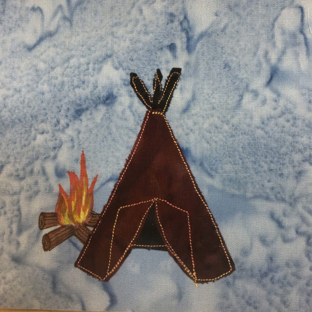 This quilt block shows a teepee and a little campfire against a blue salmon-patterned background. The teepee is in a cone shape and made out of very dark brown fabric, with darker sticks and inside. This fabric, including the tent flaps, are outlined with yellow and orange stitching. Next to the teepee is a campfire drawn with pen. The logs are round, and brown, while the fire is yellow and orange flames.