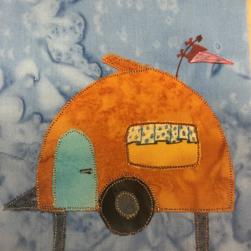 This quilt block shows an orange little rounded camper trailer. This trailer is on gray stilts at the moment, though it does have a big black wheels in the bottom center (with a dary gray hubcap piece of fabric). On the left side is a teal door, and on the right side is a window with little polka dot drapes at the top. Above that, on the dome of the orange camper is an antenna with a red little flag attached to it. This is all against a light blue salmon-patterned background.