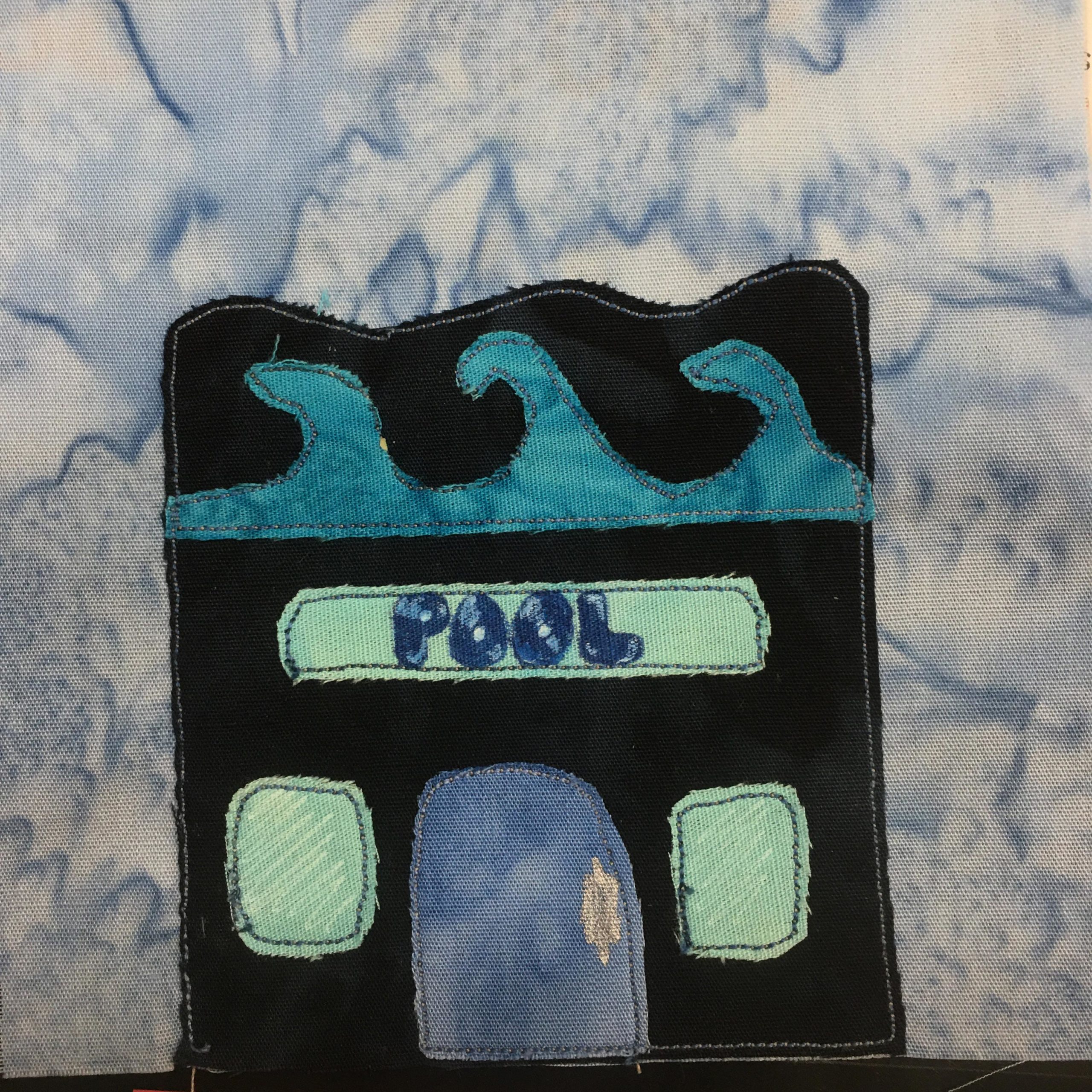 This quilt block shows a building with a bumpy roofline. Underneath the wavey roofline is actual waves made out of blue fabric. The building itself is completely black, except for the windows, sign, and door. The sign under the blue waves is turquoise with blue letters written in pen,