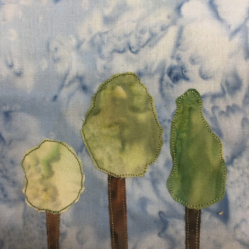 This quilt block shows three trees in a row with various foliage. Each tree has a brown trunk with green stitching outlining it. The foliage is indicated by pieces of green fabric, also with green stitching outlining them. One tree is short with a round foliage, the next is medium with an oval foliage, and the next is tall, but with a short trunk, with a cone-shaped foliage. This is all against a blue salmon-patterned background.