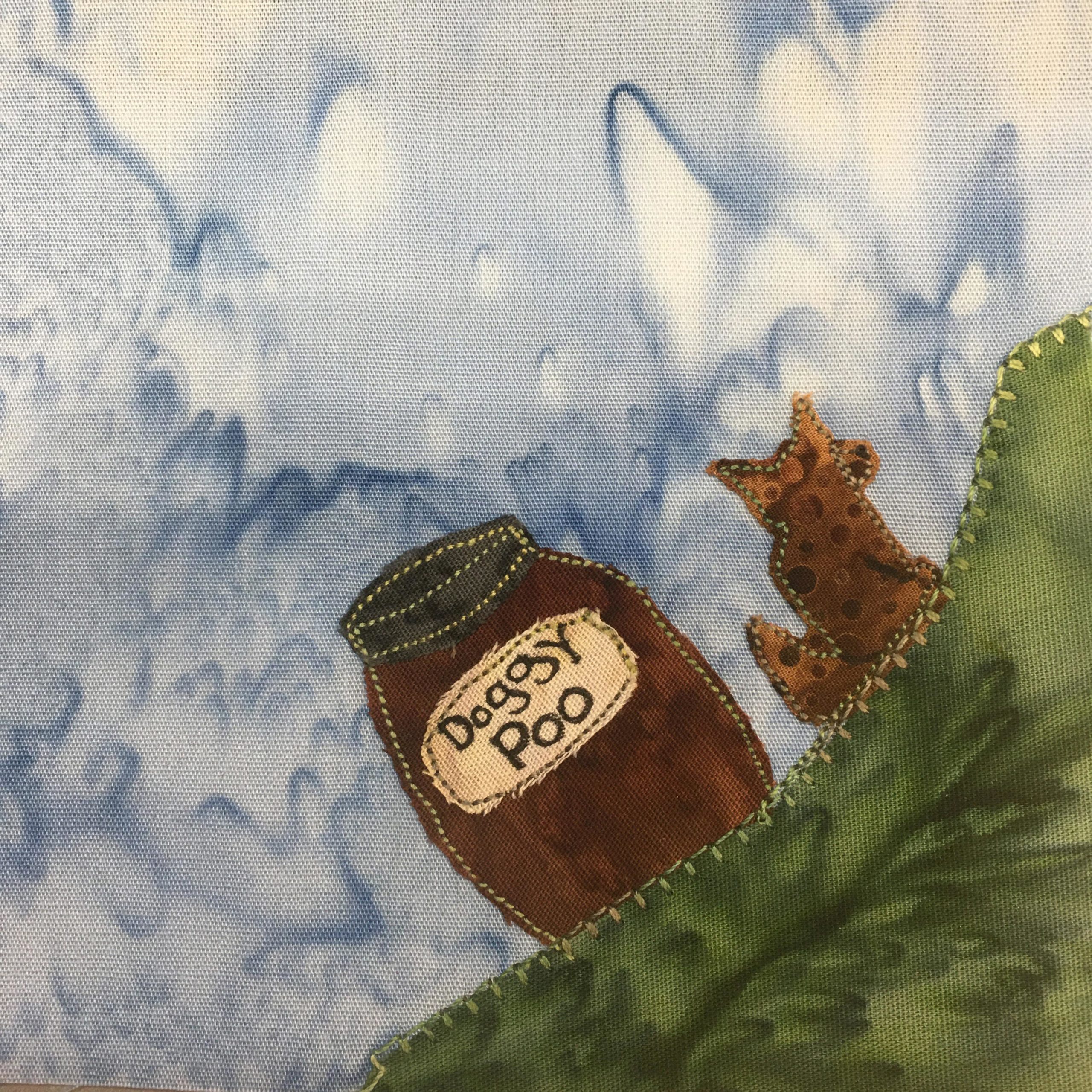This quilt block is a corner block. In the bottom right corner is a dark green patch of fabric indicating grass with a nice green blanket stitch. On top of the grass, pointing towards the center of the block is a brown polka-dot duotone dog shape (representing a dog) with green stitching around it's border. Next to the dog is a brown bin with a gray top that reads, "Dogsy poo." The bin is outlined with the same green stitching.