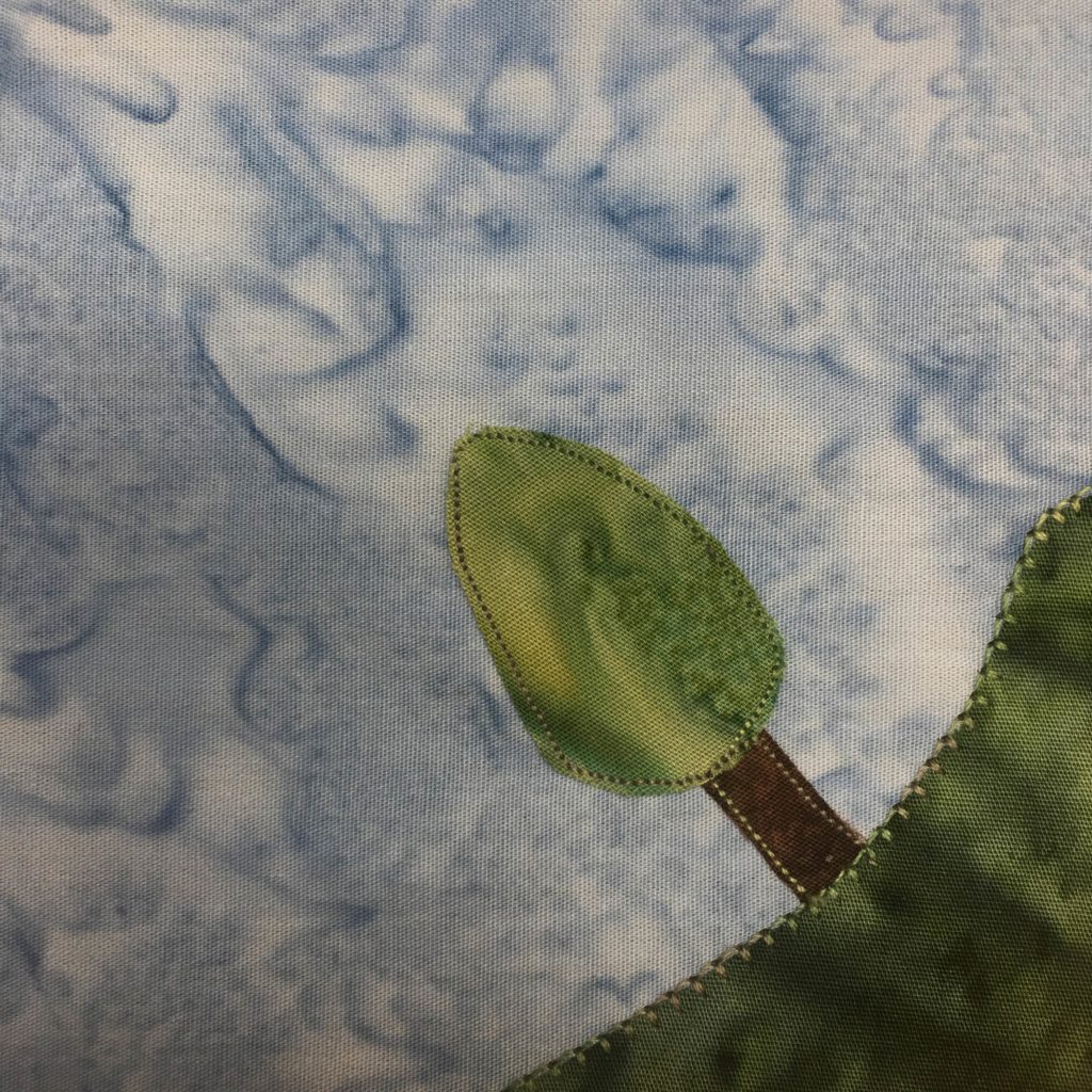 This quilt block is a corner block with a green section of fabric in the bottom right. This piece of fabric has a blanket stitching with equally deep green thread. Coming out of the green section and pointing towards the center is a tree. The trunk is dark brown, and the foliage is a bit cone shaped. It's the same green as the grass, and each piece is outlined in the same green stitching.