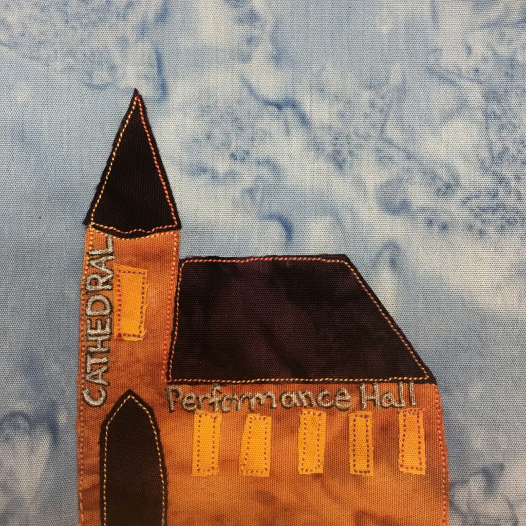 This quilt block shows a very orange church building. The steeple of the church building is tall and has a triangular top. Next to it is the long roof of the sanctuary. Both roofs are very dark brown, along with the door. Along the side the of the steeple written in white pen are the word,s "Cathedral" and a bright orange patch of fabric indicating a window. To the right of the rounded door are more bright orange rectangular windows and above them the words "Performance Hall"  penned in white. This is all against a blue salmon-patterned background.