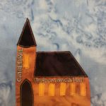 This quilt block shows a very orange church building. The steeple of the church building is tall and has a triangular top. Next to it is the long roof of the sanctuary. Both roofs are very dark brown, along with the door. Along the side the of the steeple written in white pen are the word,s "Cathedral" and a bright orange patch of fabric indicating a window. To the right of the rounded door are more bright orange rectangular windows and above them the words "Performance Hall" penned in white. This is all against a blue salmon-patterned background.