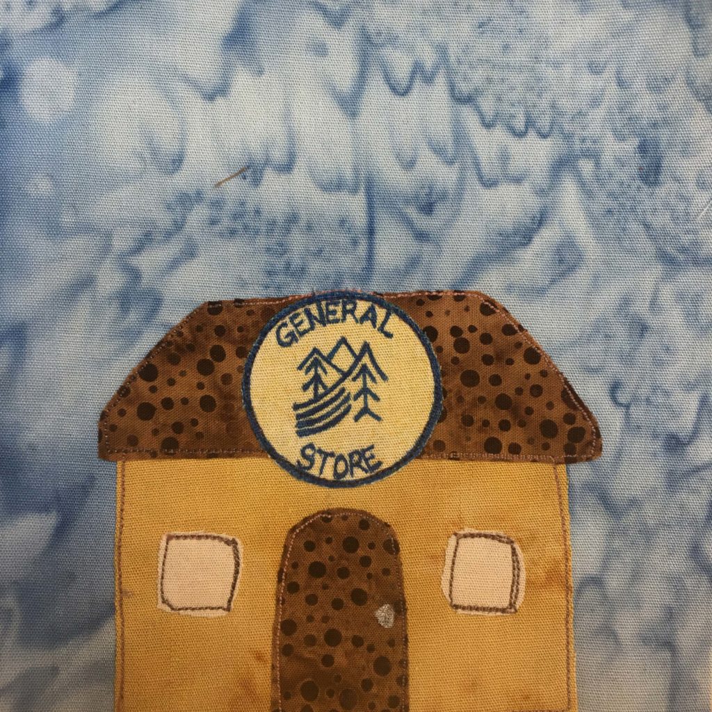 This quilt block shows a squat sqaure building with a trapezoid shaped roof. The roof and the door is made up of a duotone brown polka-dotted fabric. The building itself is an orange-tan color, with lighter tan square windows on either side of the door. The windows are outlined in a brown stitch, about the color of the roof. On the roof is a big sign that looked like a badge. It's a blue circle with a light yellow interior fabric. On the fabric written in blue pen is a logo showing trees, mountains, and a river, and the words, "General Store" along the outside circumference.