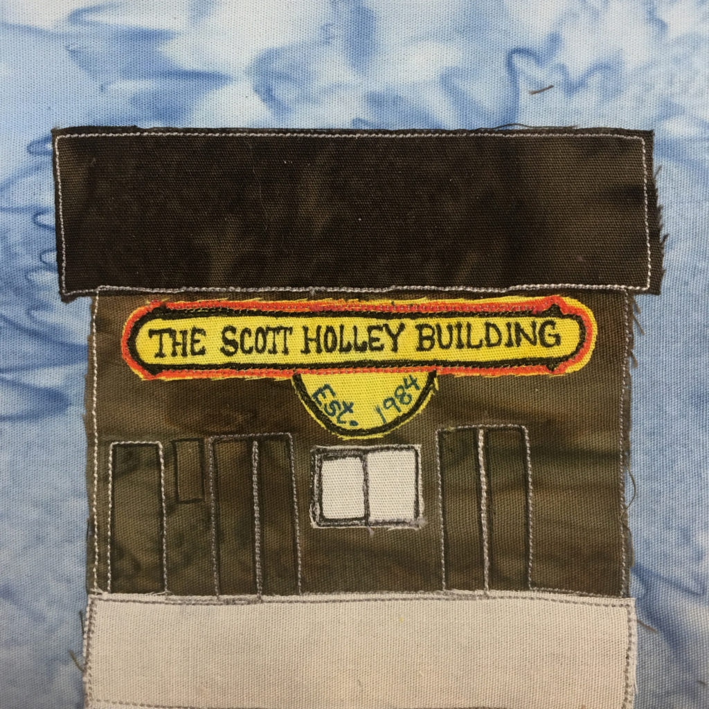 This quilt block shows a large dark gray building that takes up most of the block. The roof is the darkest gray and is square and flat. Underneath that is a very big sign that's yellow. It reads,