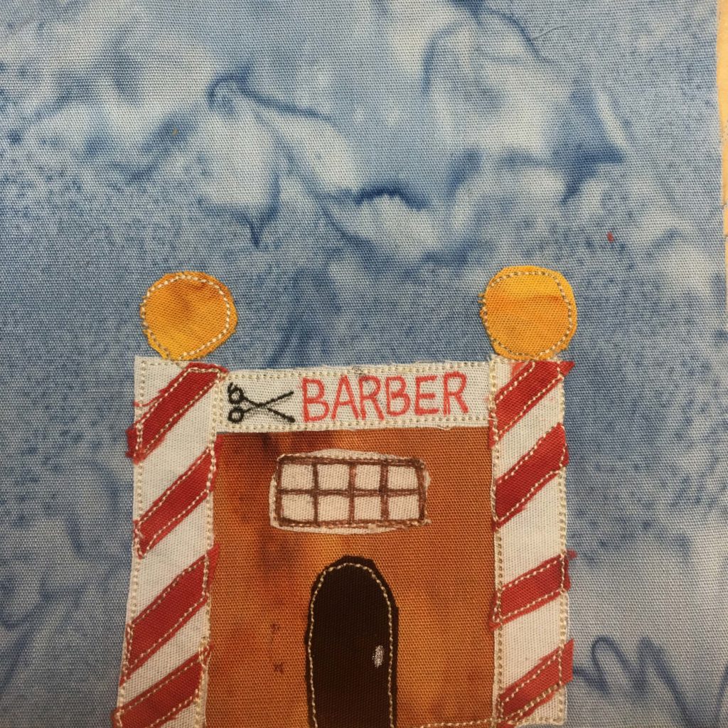 This quilt block shows an oddly shaped building. It's square but either side of it is a candy-striped barber pole with a gold ball on top. Along the top or roof is a white stripe that has scissors drawn on it and the word "Barber" in red. Underneath that is a grid window, really a patch of light cloth with squares drawn in brown in pen. It's an orange building otherwise, with a dark brown rounded door. This is all against a blue salmon-patterned background.