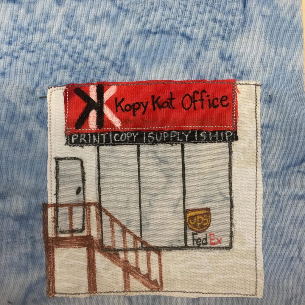 This quilt block shows a square building with a sign on it and a wooden stairway up to a door. The building is white with black outlines showing the door and the windows. Inside the big window that's divided into three panes, there are the signs for UPS and FedEx penned in. The wooden staircase is brown and penned in to go up to the door. Above everything, hanging off the flat roof is a red sign. There's a black K backwards on top of a white K, and the words next to it are