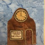 This quilt block shows a clock repair shop. it's shaped like an old mantel clock but with a bigger base, which is the rest of the building. Theere is a big clock face pointing 12:20 at the top of the roof. The building is all brown, like a wooden mantel clock. There is a window on the left and in it in brown letters reads,