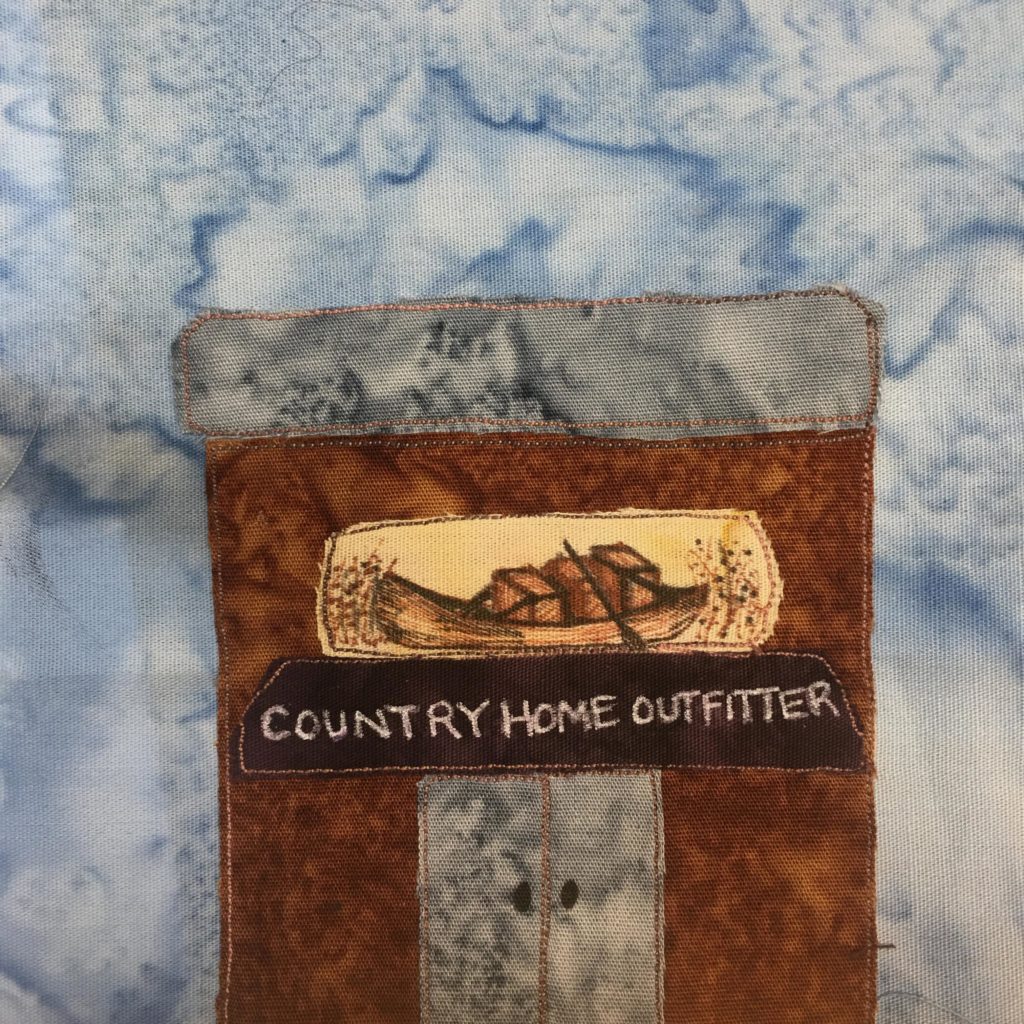 This quilt block shows a square building. The roof is gray and flat. Underneath that is a brown building with gray double doors. There is a very detailed picture of a long boat drawn in pen on a yellow piece of fabric above the door. Below that, still above the door, is a dark brown sign with white letters that reads,