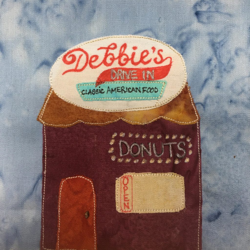 This quilt block shows a square building with a big round sign on top. The roof has a curvy bottom, and is a light brown. The big oval white sign on top has the word "Debbie's" in cursive and red. There's a red tail coming from the S and on it it says underneath, "Drive In" Below that is a light blue trapezoid that says, "Classic American Food." The building itself is a deep maroon. In lights, or dots, is the word, "Donuts." Below that is a yellow window, with a red "Open" sign vertical. Then there is a brighter red door to the left.