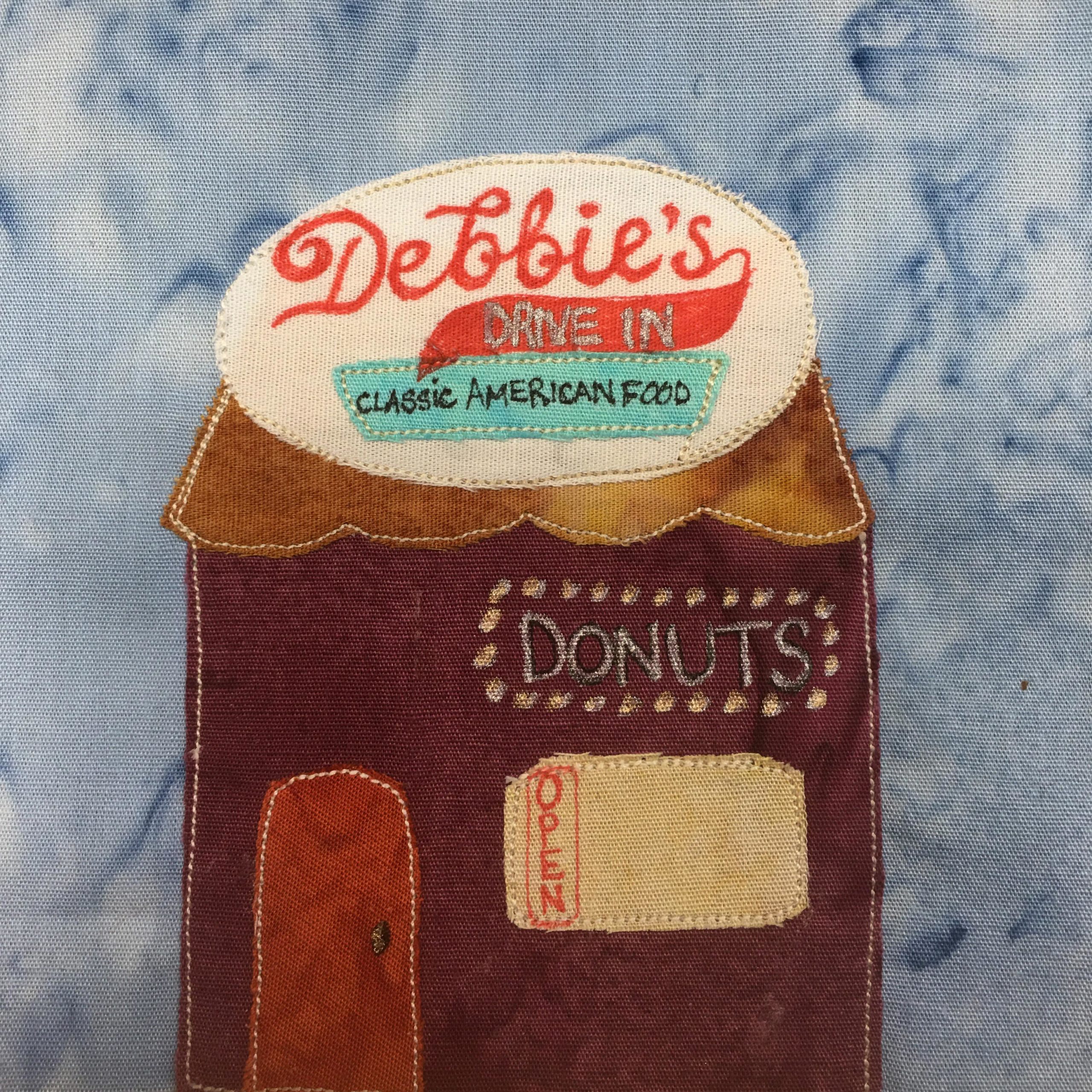 This quilt block shows a square building with a big round sign on top. The roof has a curvy bottom, and is a light brown. The big oval white sign on top has the word "Debbie's" in cursive and red. There's a red tail coming from the S and on it it says underneath, "Drive In" Below that is a light blue trapezoid that says, "Classic American Food." The building itself is a deep maroon. In lights, or dots, is the word, "Donuts." Below that is a yellow window, with a red "Open" sign vertical. Then there is a brighter red door to the left.