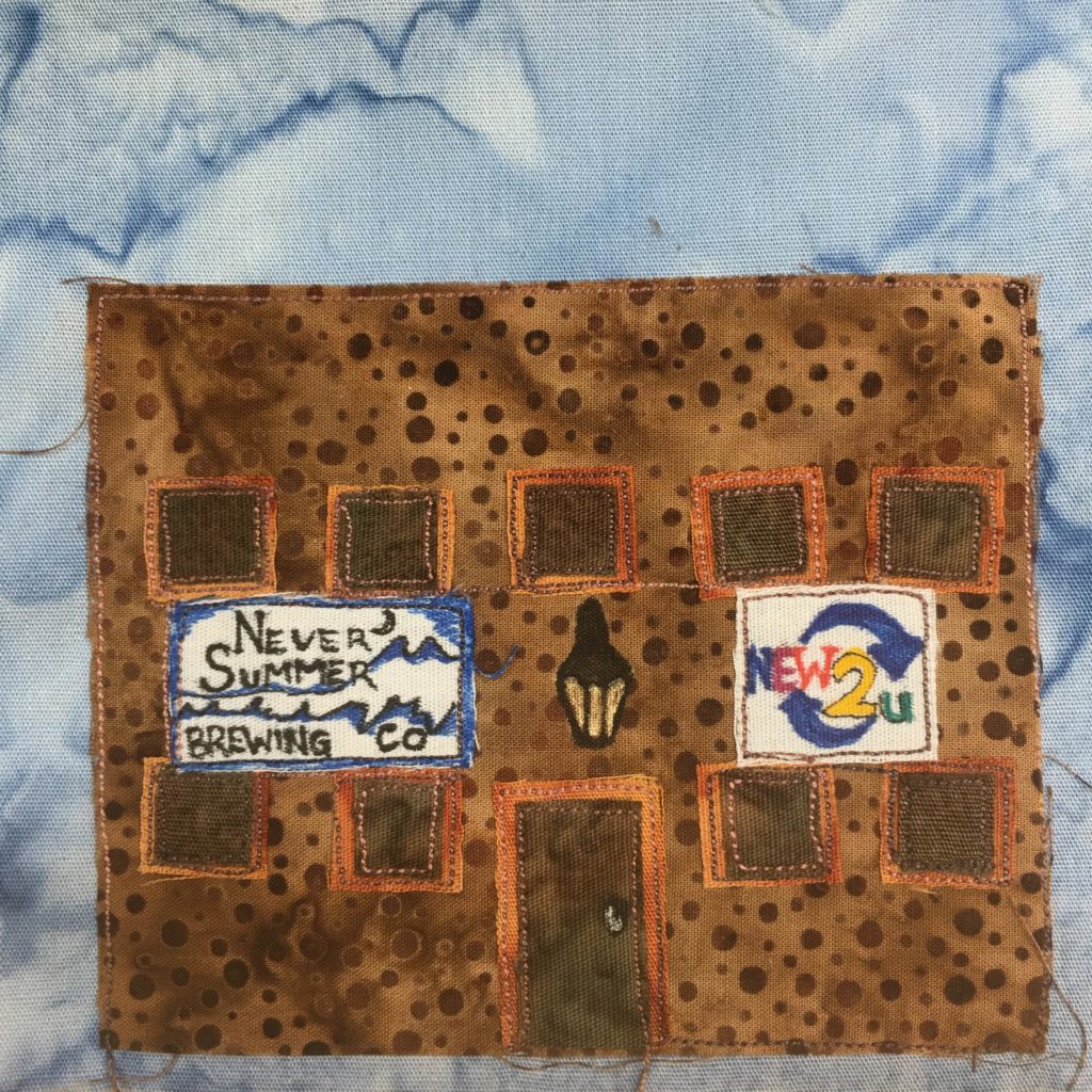 This quilt block shows a large just about square building. The main building material is a single large duotone polka dotted fabric (the dots being scattered and random). There are many smaller square windows outlined with an orange fabric, while inside they are a solid brown. They're each outlined by stitching. There is a light fixture above the middle door drawn in pen. The door is in the middle and is also outlined in an orange fabric. There are two signs, each detailed and colorful. On the left is a blue, black, and white sign with mountains on it and thew words,
