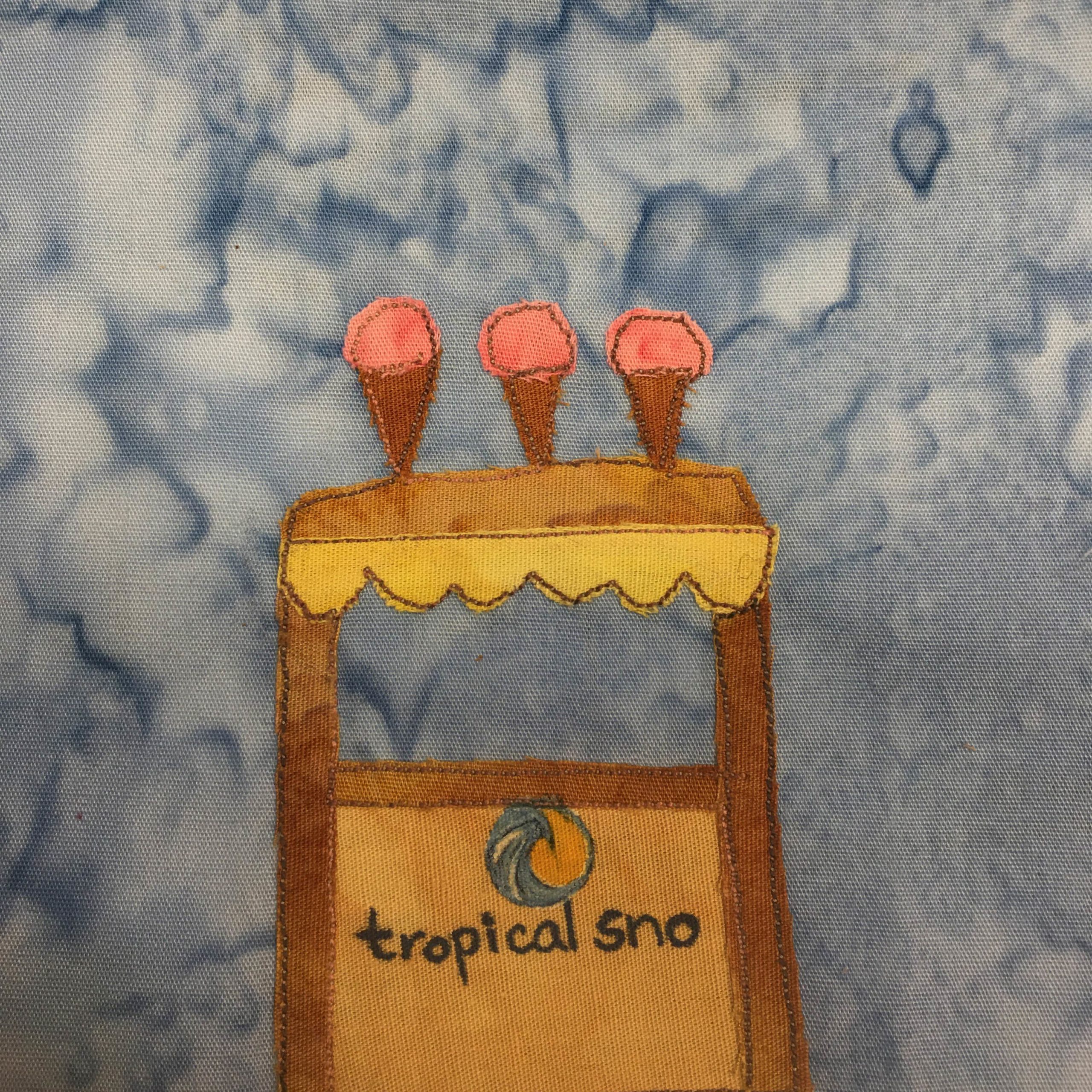 This quilt block shows a seasonal snocone stand. The top of the snocone stand has giant fake snocones coming out the top. They are pink and look like ice cream cones. Below that is a yellow canopy with a frill at the end. Then there are boards forming an H that are brown. Below the middle board is the front of the stand, which is a darker yellow than the frill. On the front is a picture of a wave in a circle, and underneath that the words in black pen, "Tropical Sno"