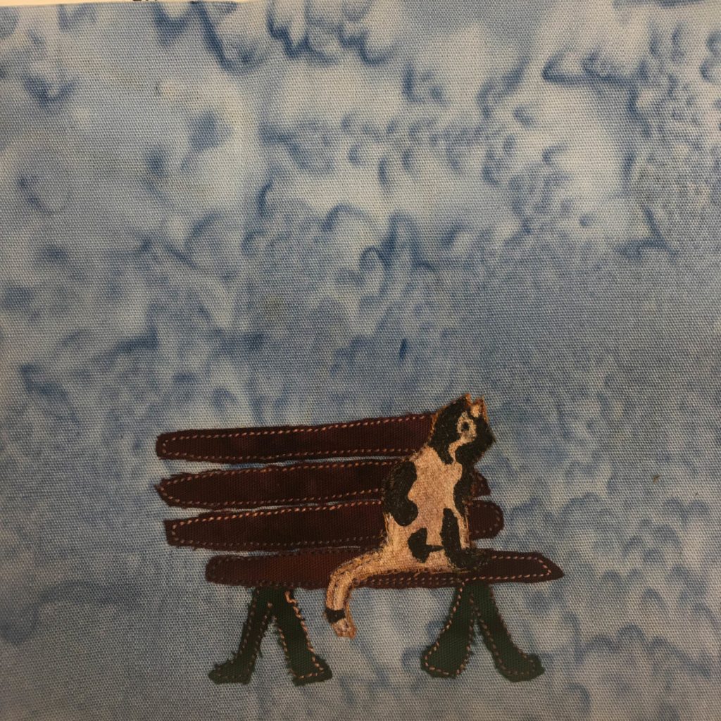 This quilt block shows a dark brown and maroon bench with a cat sitting on it. The cat is a light tan cream color with black splotches. The bench sits on two black v-shaped stands. The slats are horizontal and outlined with stitching. This is all against a blue background.