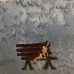 This quilt block shows a dark brown and maroon bench with a cat sitting on it. The cat is a light tan cream color with black splotches. The bench sits on two black v-shaped stands. The slats are horizontal and outlined with stitching. This is all against a blue background.