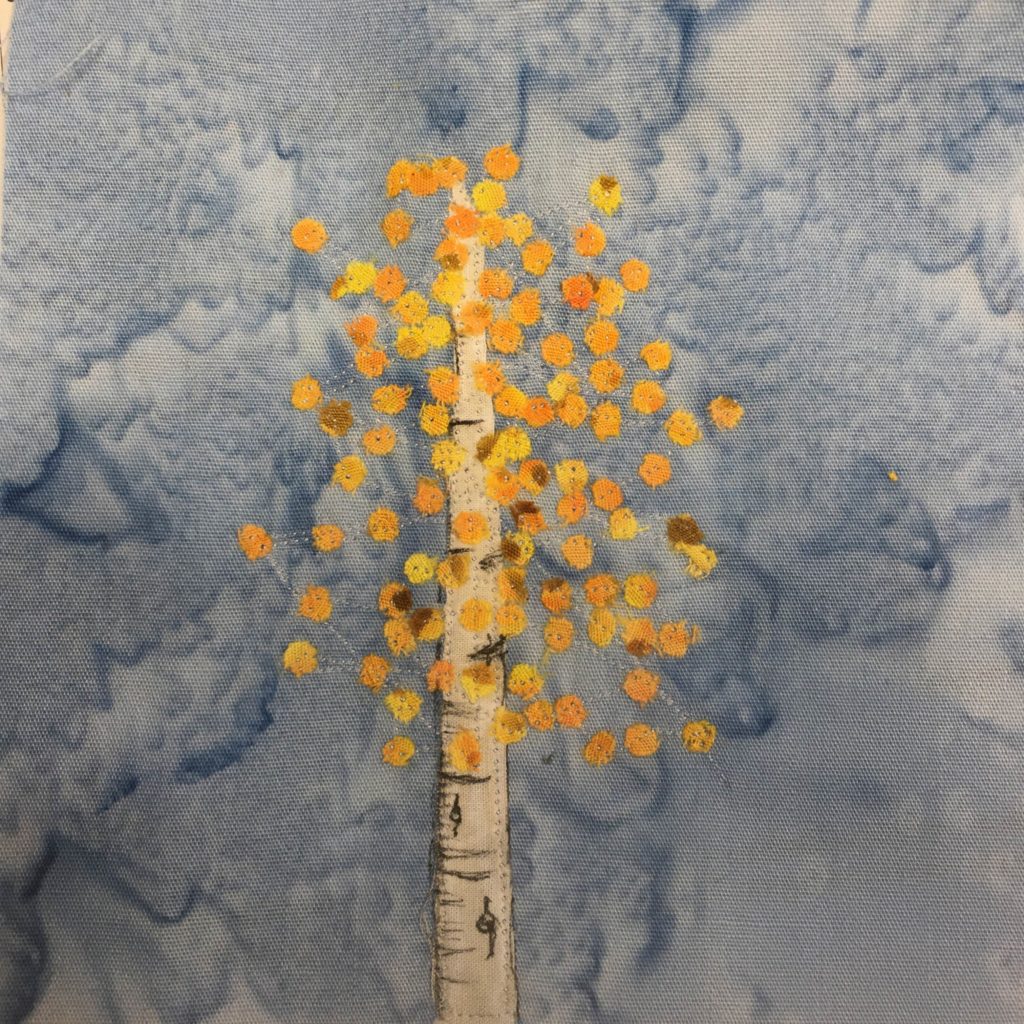This quilt block shows an aspen tree. The tree trunk is a cream colored fabric stick with darker lines drawn in to give it the texture of an aspen tree. The leaves are numerous little yellow and orange spots of fabric, all attached with invisible thread. They appear as if they are floating around the aspen tree trunk, and that they are turning for autumn.