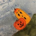 This quilt block is a corner block. There is a dark green grass patch in the bottom right of the square. On that grass patch are two jack-o-lanterns. The one in front is the traditional bright orange pumpkin with a spooky black face drawn on it with pen. There is also some thread or pen indicating a bit of a vine on it. Behind it is a tall squash that's more yellow. On it is a cuter spooky face with more rounded features. The top of this squash has some orange on it. This is all against a blue salmon-patterned background.