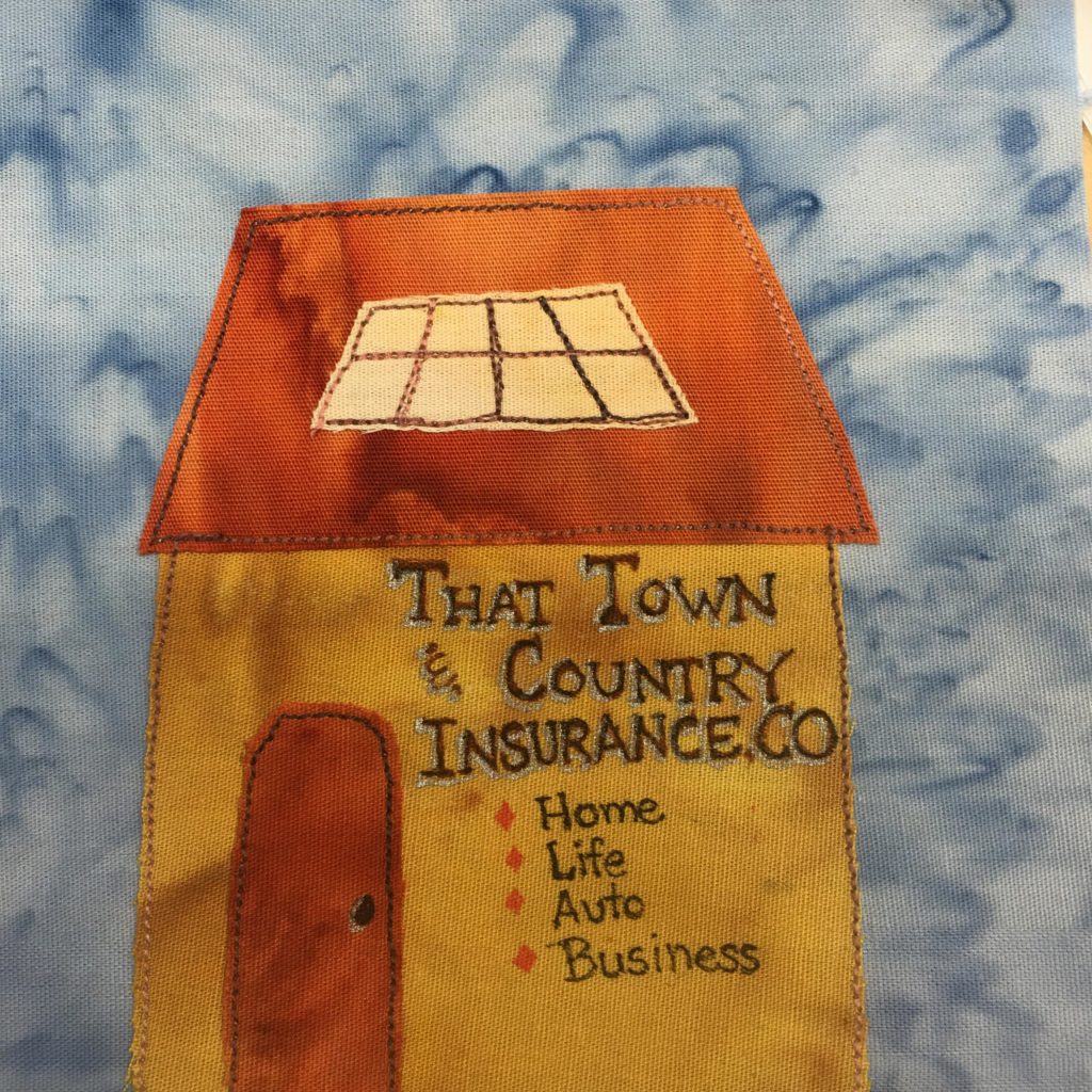This quilt block shows a tall building with a large skylight. The roof is a large orange trapezoid. On the center of the roof is a yellow piece of fabric that is also in the same shape as the roof. There are threads that separate the panes of glass in the skylight. Below the large roof Is a dark yellow/gold main body. On that main body is large writing with a white outline that reads,