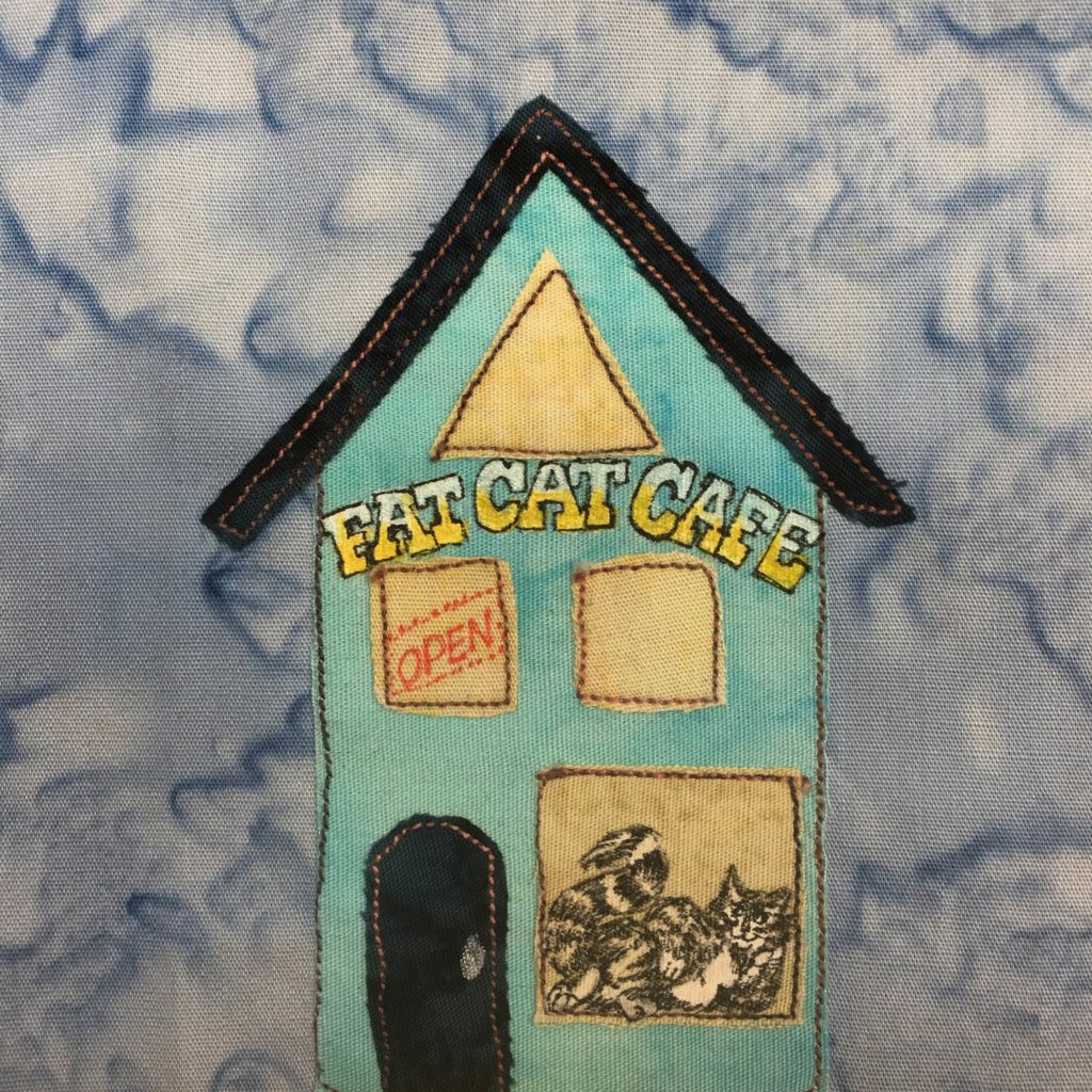 This quilt block shows a tall blue building with an angled roof. Below the dark blue roof is a triangle window, in the same shape as the peak of the roof. Below the pink yellow window on the light blue building is a sign. The lettering is bold and has a gradient from bottom to top from yellow to even lighter blue. It says, "FAT CAT CAFE." Below that are two yellow windows, the left one having a red open sign. Finally on the ground level is a large window with a very stylish cat in a very stylish pose like out of a cartoon. It's striped and black and white. To the left of the cat logo window is a dark blue door the same shade as the roof. Both the door and the roof have yellow/gold stitching outlining them.