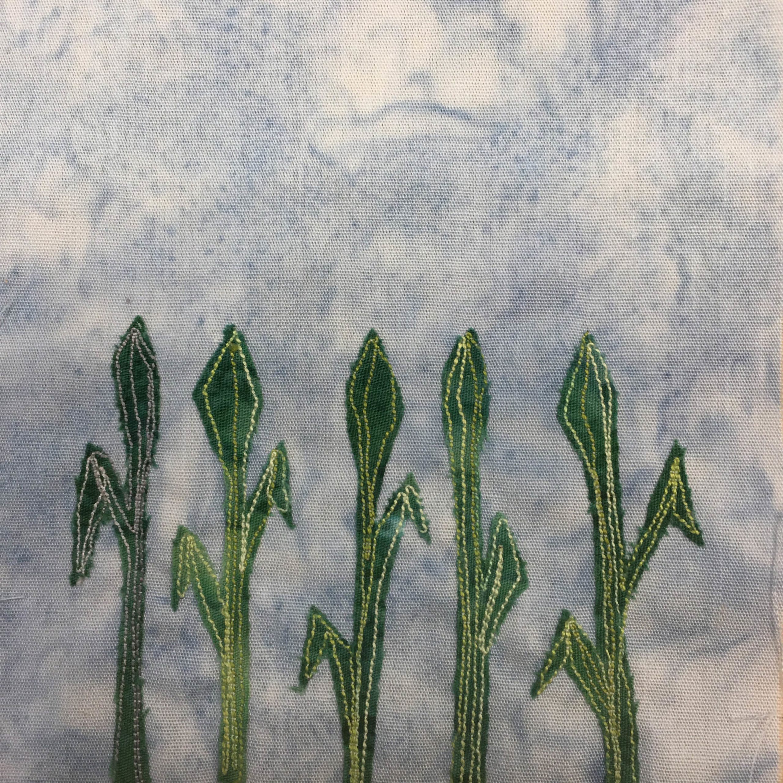 This quilt block is fairly simple. It's five plants of corn against a very light blue background. Each plant comes up from the bottom and stops at about the same height, midway up. Each plant has two leaves coming off of it except the left which has one. At the top of each piece of green fabric is a bulb that's covered in leaves. The texture of the corn plant is indicated by using parallel rows of yellow stitching up and down the corn.