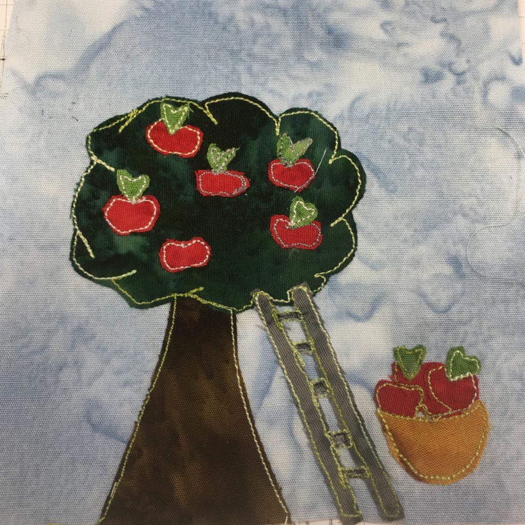 This quilt block shows an apple tree with apples in it. The trunk is a dark brown and very wide, tapering up towards the foliage. In the dark green of the greenery are red pieces of fabric with bright green tops that are apples. The foliage is outlined by a bright green stitching. Propped up against the tree is a silver ladder, and nexto the silver ladder is a satchel that's tan with several apples of the same kind inside.
