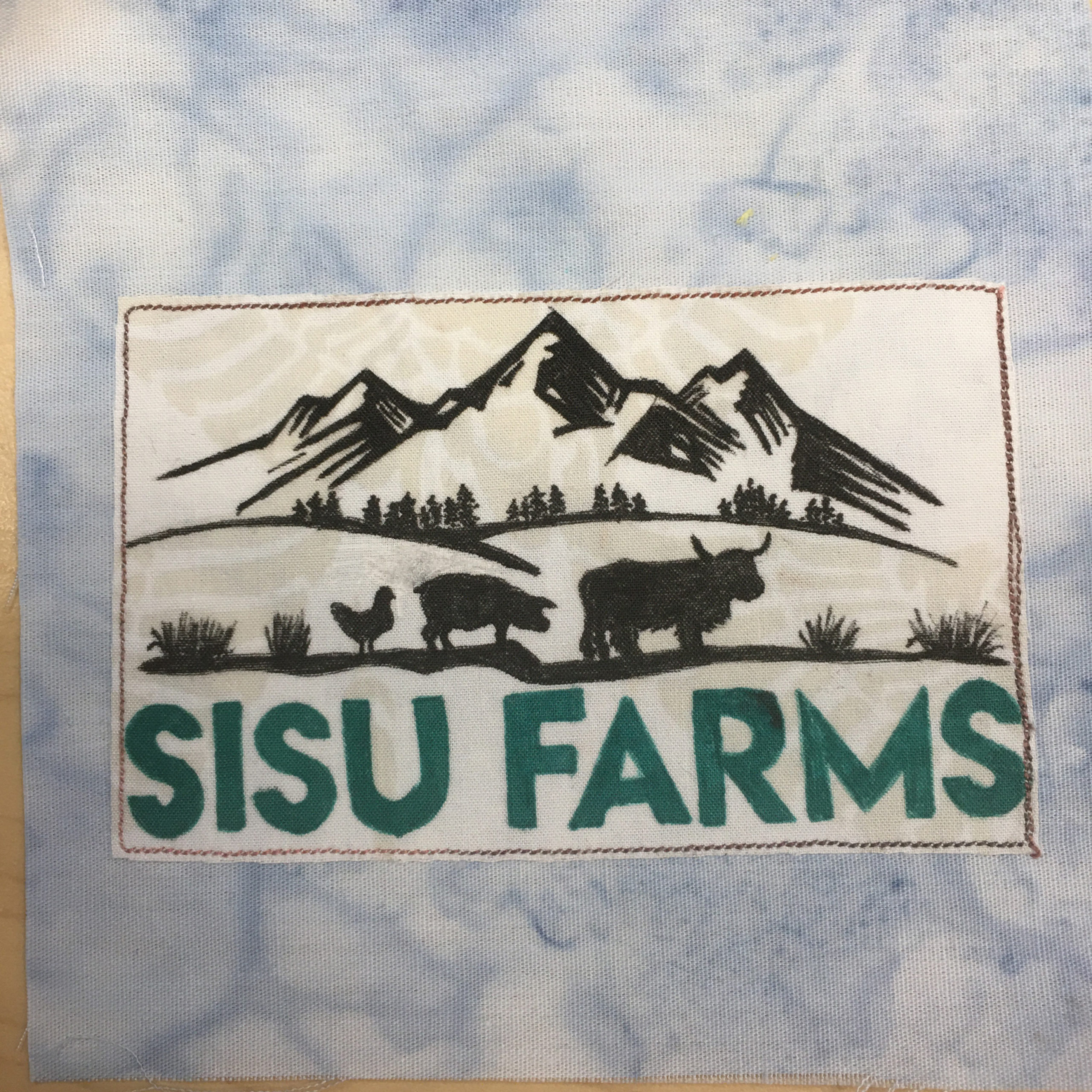 This quilt block shows a white square in the middle on top of a salmon-patterned blue background. The square in the middle has a pen drawing on it. The drawing shows a mountain range at the top, with fields and trees below. O the last line of ground are a chicken, a pig, and a highland cow. Below this elaborate black and white drawing are the words