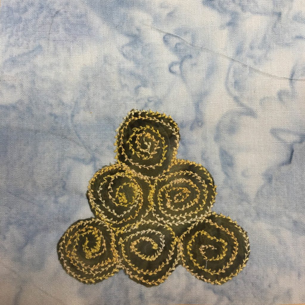 This quilt block shows a blue background with six round bales of hay in the bottom center. The bales of hay are actually round, and we are looking at them from the side. Each bale is a green piece of fabric with a bright yellow leaf-like stitched spiral coming out from the center quite dramatically. They are placed in a pyramid pile with three rolls at the bottom.
