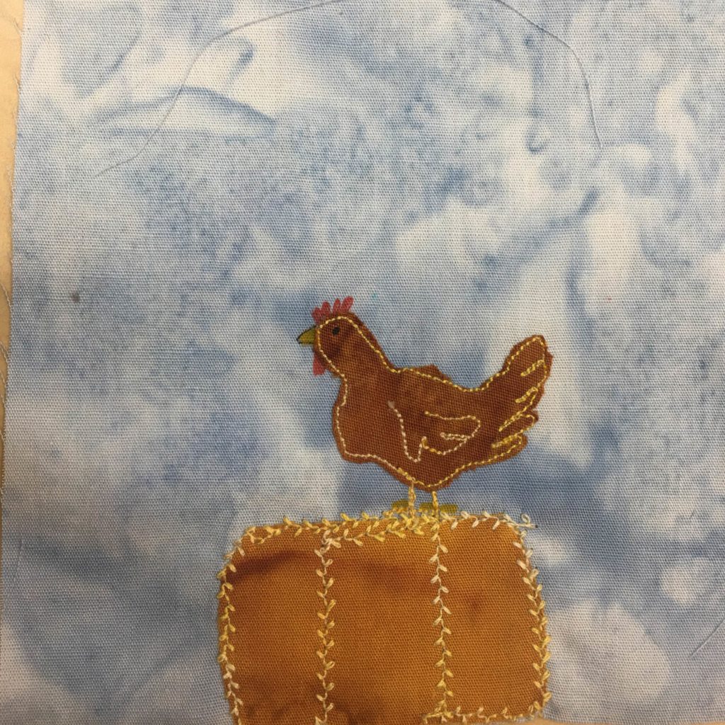 Against a blue background we can see a bale of hay, and on top of that bale is a chicken facing to the left. The chicken is a darker brown fabric with yellow stitching on it outlining its shape and forming a wing. At the head of the chicken is a beak and eye drawn in yellow, black, and red pen. The bale of hay is a square orange block with a special leaflike yellow stitching going around it and up th middle in sections.