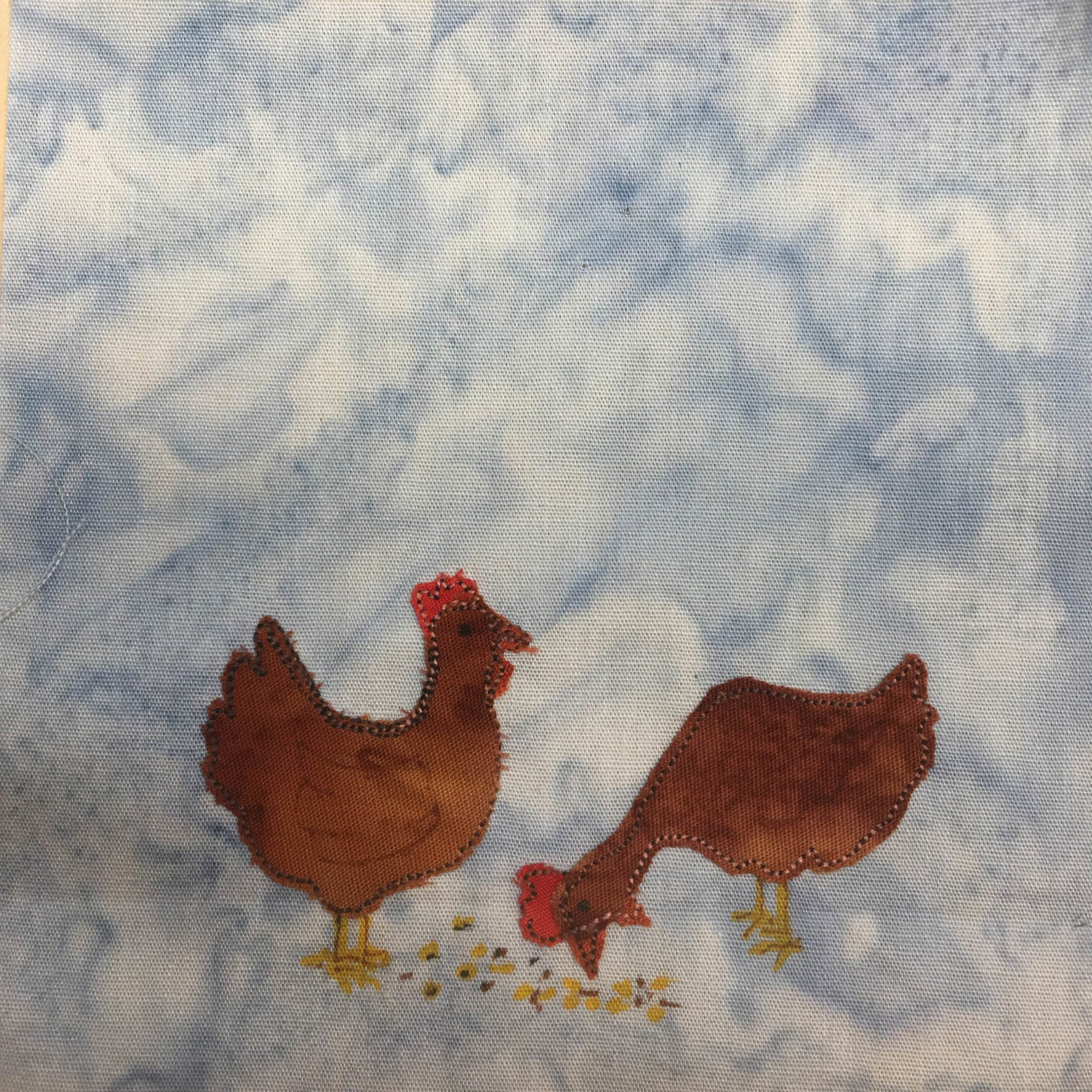 This quilt block shows two chickens facing each other. One chicken is pecking on the ground where there are some yellow dots and brown dots indicating scratch. The other chicken is upright looking onwards. Each chicken is a brown piece of fabric with a light orange and black stitched outline. The chicken's crests are small rec pieces of fabric with similar stitching. The feet and beak are drawn in with pen. This is all against a light blue salmon-patterned background.