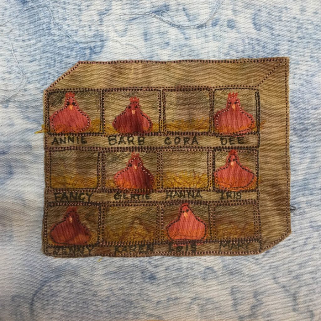 This quilt block shows a brown piece of fabric made to look like a big rectangular box on its side with demarcated boxes in pen in it. In each box is either yellow hay, or a light rust colored hen sitting on eggs looking at the viewer. This is against a blue splotch background fabric.