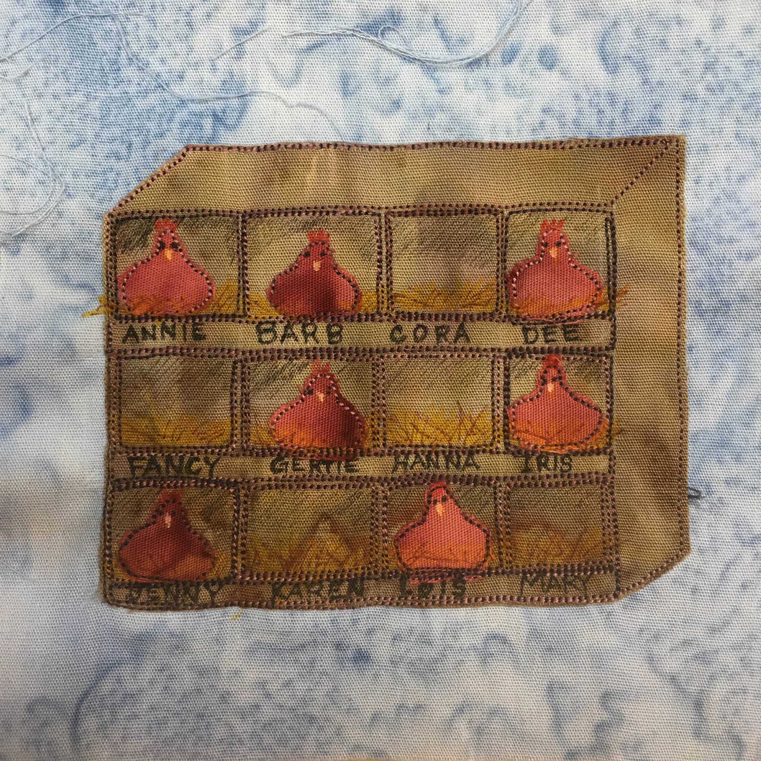 This quilt block shows a brown piece of fabric made to look like a big rectangular box on its side with demarcated boxes in pen in it. In each box is either yellow hay, or a light rust colored hen sitting on eggs looking at the viewer. This is against a blue splotch background fabric.