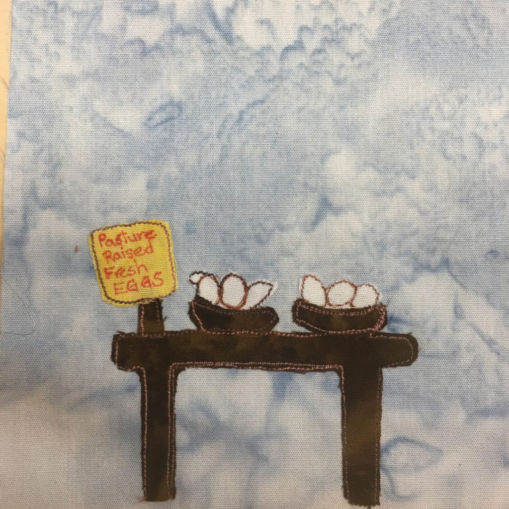 This quilt block shows a table with bowls on it. In the bowls are white eggs. There is a sign next to the two bowls holding the eggs which is yellow. There is red lettering on it that says,