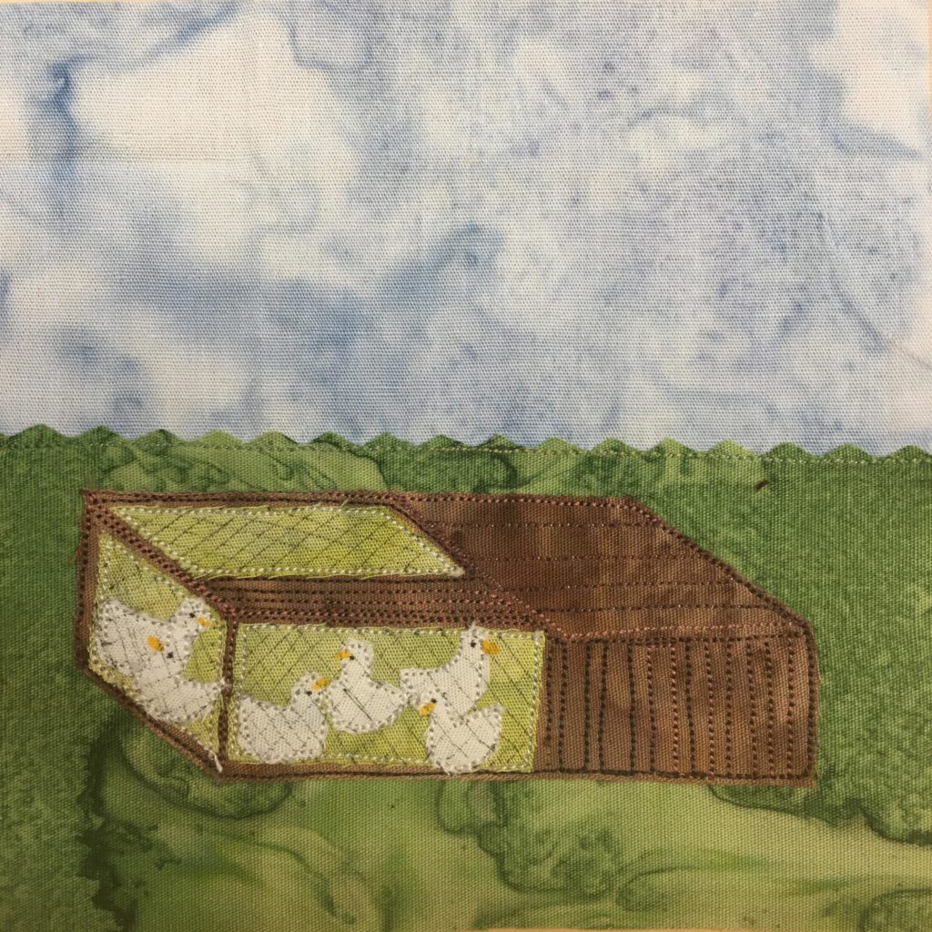 This quilt block shows a short box on a field of grass with white hens inside. The bottom half of the block is a green salmon-patterned fabric, while the top half is a light blue salmon-patterned baground. In the middle of the green fabric are brown pieces of fabric with brown stitching that show a squat structure. Half of the structure is covered, the other half is lined with chicken wire. Inside the chicken wire are white chickens, each it's own piece of fabric.