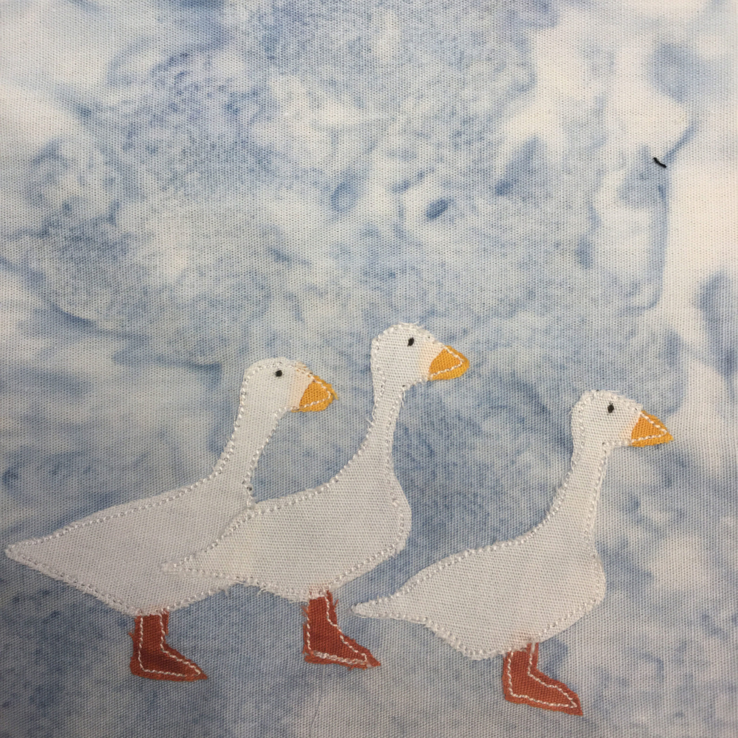 This quilt block shows three large white birds against a light blue salmon-patterned background. Each bird is actually a goose. Each goose, all facing right, is made up of a darker orange fabric for its feet, a white piece ofgabric, and a little yellow piece of fabric for its beak. Each of these are outlined in white stitching.