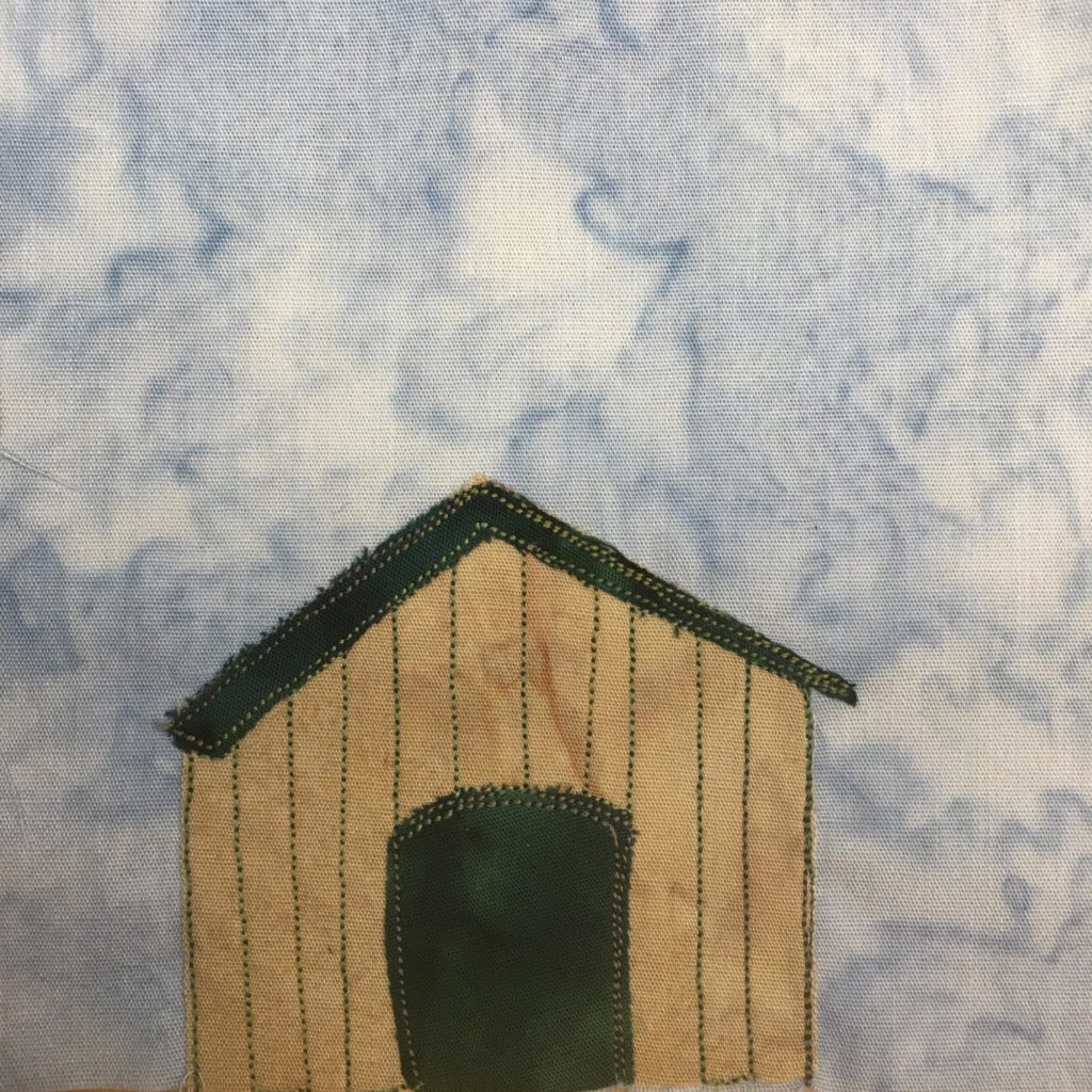 This quilt block shows a tan squat building with a dark green thin angled roof. The opening, much like a dog house, is in the middle and is also dark green. The dark green pieces of fabric are stitched with a lighter green stitching. There are also lighter green stitches on the tan building to show the slats of wood. This is all against a light blue background.