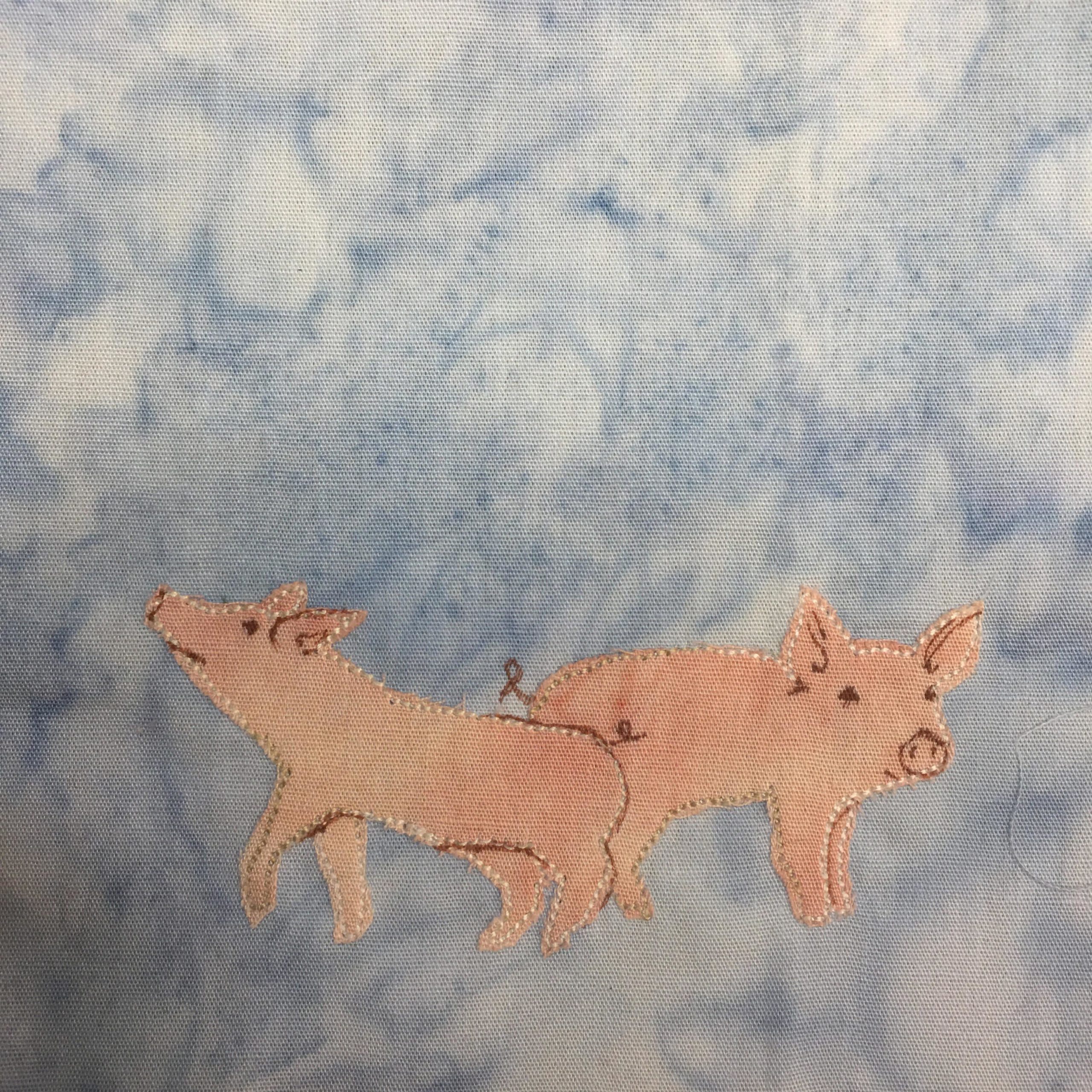 This quilt block shows two young pigs (shoats) against a blue background. The pigs themselves are a light pink color piece of fabric. There are two pigs, each facing the other direction. The left pig is sniffing the air and taking astep forward, while the other is "smiling" for the camera. Each pig is outlined by a white stitching. There are little noses and curly-q tails penned in with a brown pen.