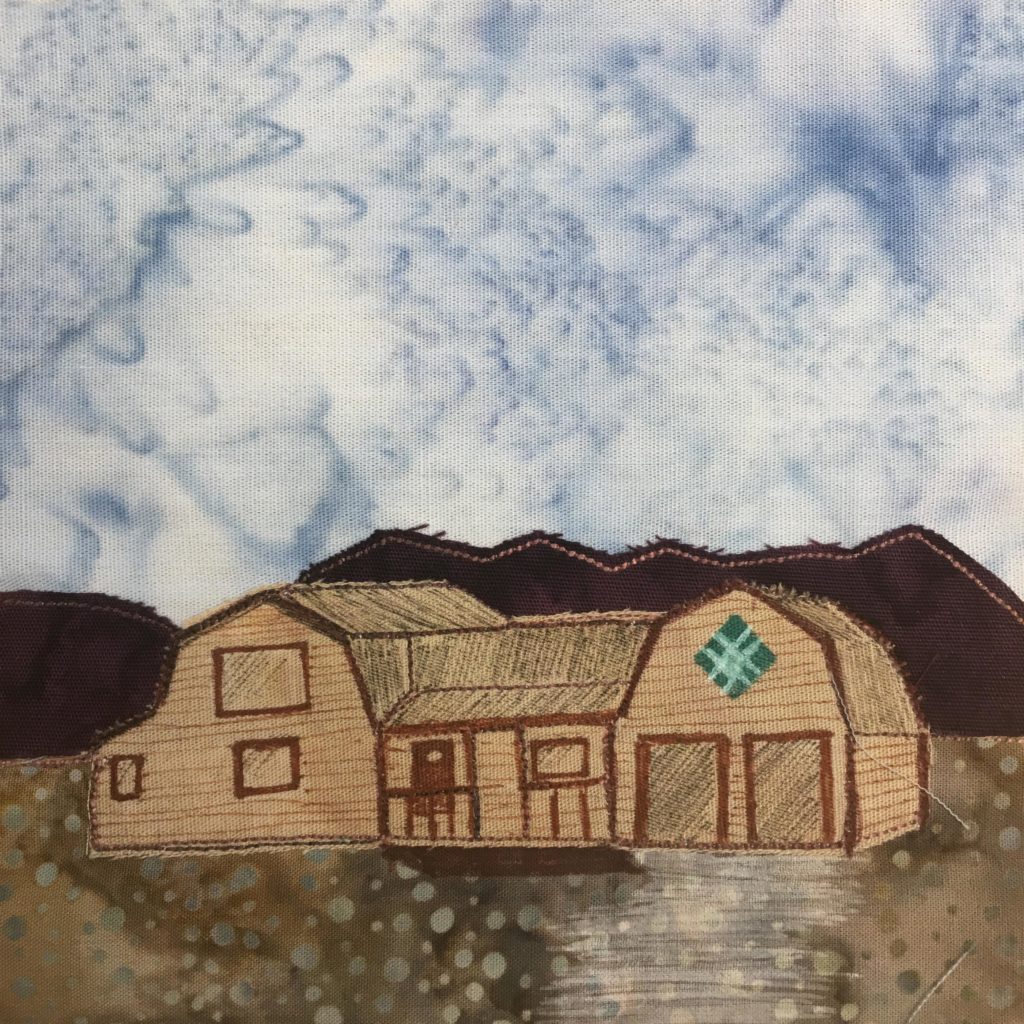This quilt block shows a house in front of a mountain range. The mountain range is a dark brown with a light brown outline stitching. The house itself looks like two barns attached to each other in the middle. It is a tan brown and has pen markings on it to indicate the metal roof, garage doors, windows and porch. There is a teal and light blue diamond shaped sign above the garage doors on the right. The ground the house is on is a polka-dotted duo-tone fabric that is a shade of brown of dirt. The sky is a light blue salmon patterned background.