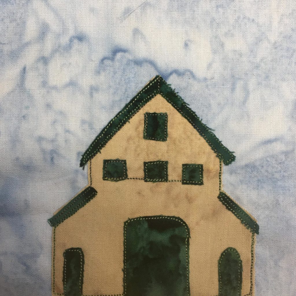 This quilt block shows a multi-level barn against a light blue salmon-patterned background. This barn is shaped like a barn with a wide triangular roof, but the center protrudes further up with a large two-story loft. The two sides are lower. The barn itself is a tan fabric with dark green windows, roofs, and doors. The dark green patches of fabric are outlined by a light green stitching. There is a small door on the left side, a huge door in the middle, and a small door on the right side. The loft has three lower square windows and one higher square window in a triangle formation.