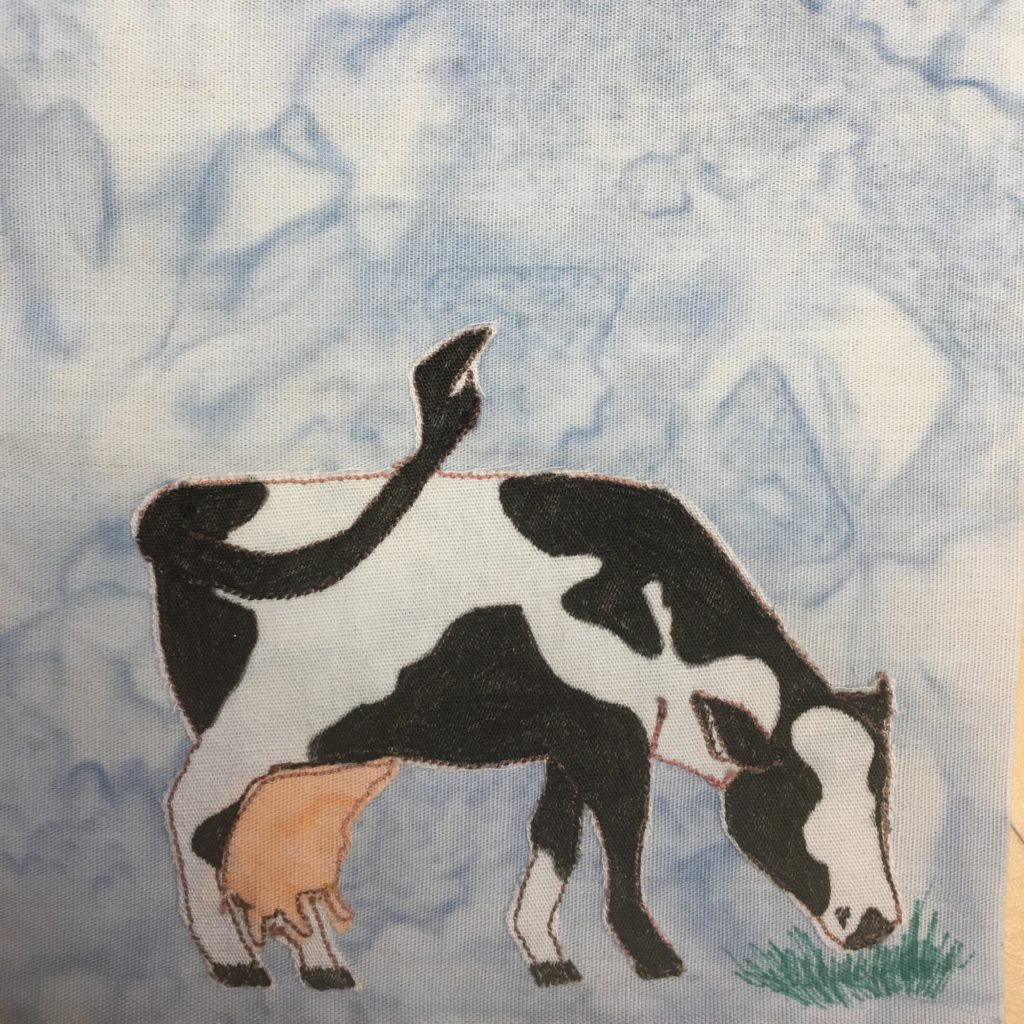 This quilt block shows your stereotypical black and white milking cow. The cow is made out of white fabric with black splotches. The udder below, which is fairly large, is made of a light pinkish fabric. Both are outlined by a very thing stitch of orange thread. The cow is facing to the right with its head down grazing on some grass drawn in with pen. The tail of the Holstein cow is long and black with a tuft at the end. It's in full swing from behind the cow.